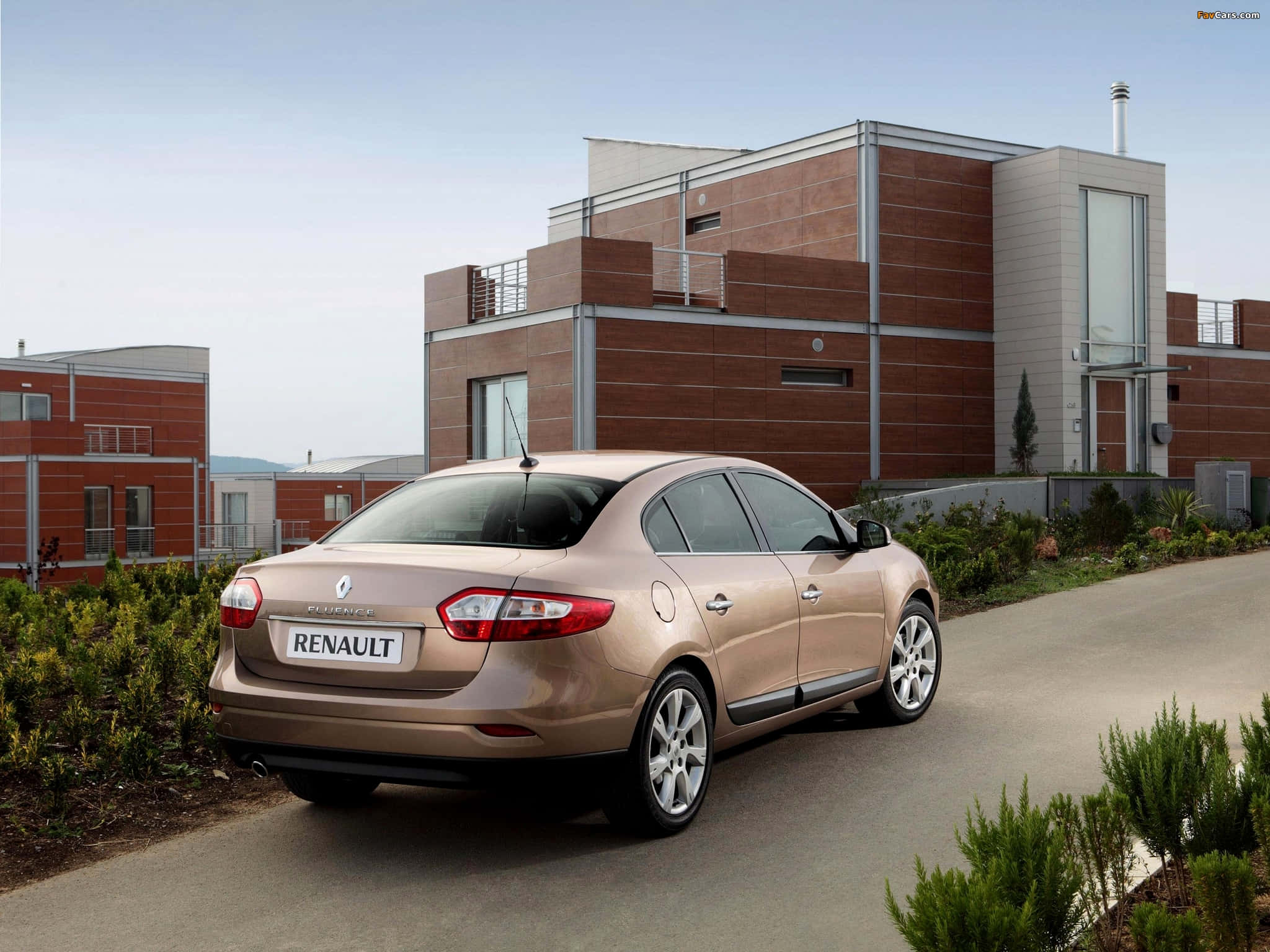 Renault Fluence 桌布 壁紙 — 一輛金色雷諾Laguna房車停放在鋪砌道路上，旁邊是現代磚砌建築，有綠色灌木，天空晴朗。