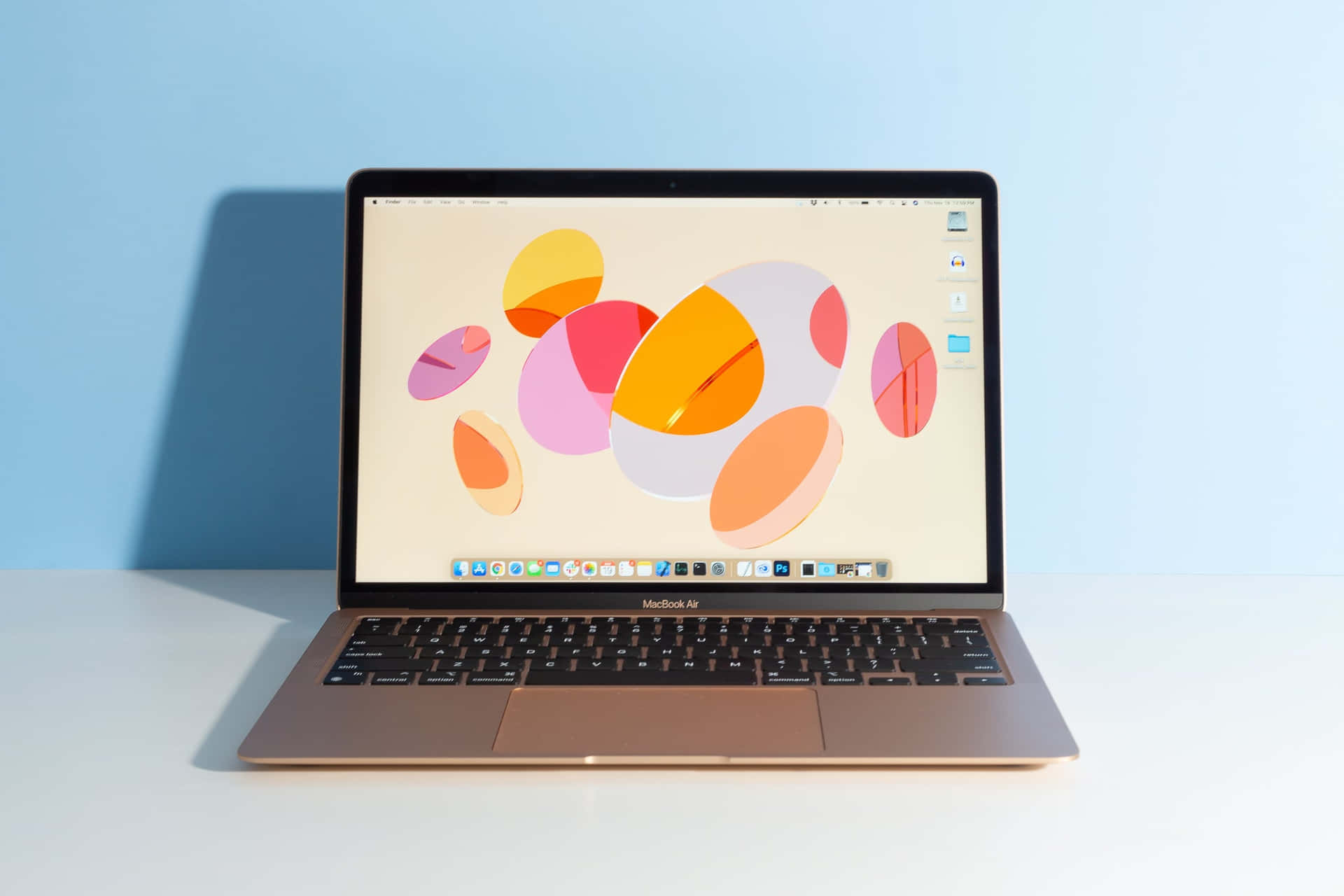 金色 MacBook Air 在白色桌子上，帶有彩色抽象桌布，背景為藍色牆壁。 — Macbook Pro 壁紙
