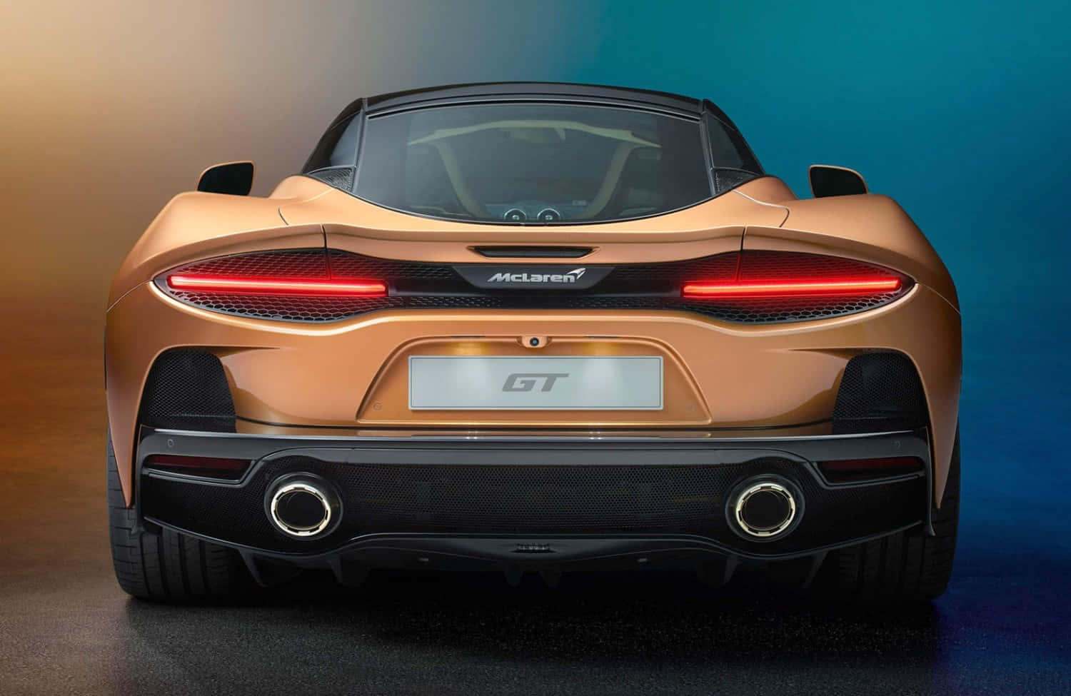 Mclaren Gt 桌布 壁紙 — 金色McLaren GT跑車的後視圖，紅色尾燈發光，背景為漸層藍色和橙色的工作室背景。