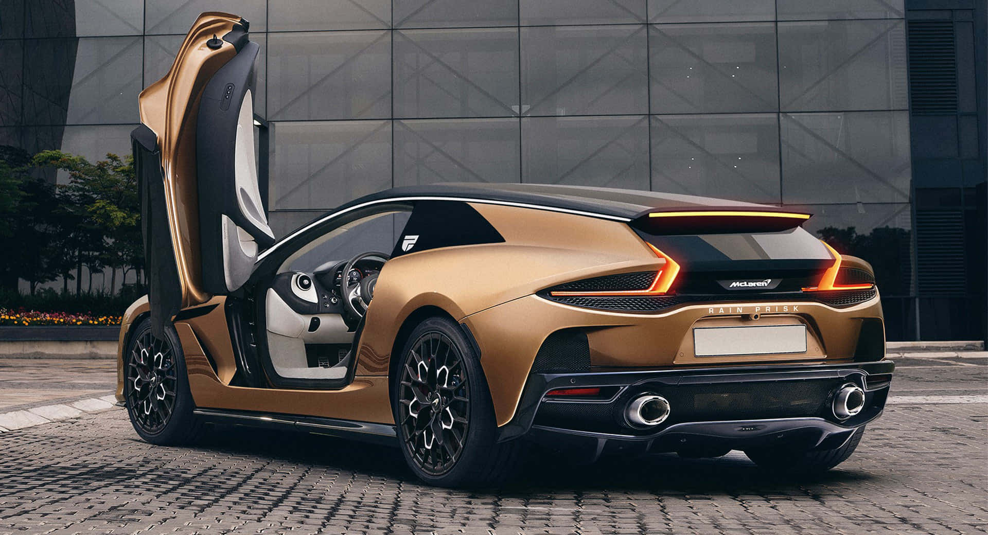 Mclaren Gt 桌布 壁紙 — 一輛金色McLaren跑車，蝴蝶門敞開，停在現代玻璃建築前的鵝卵石街道上。