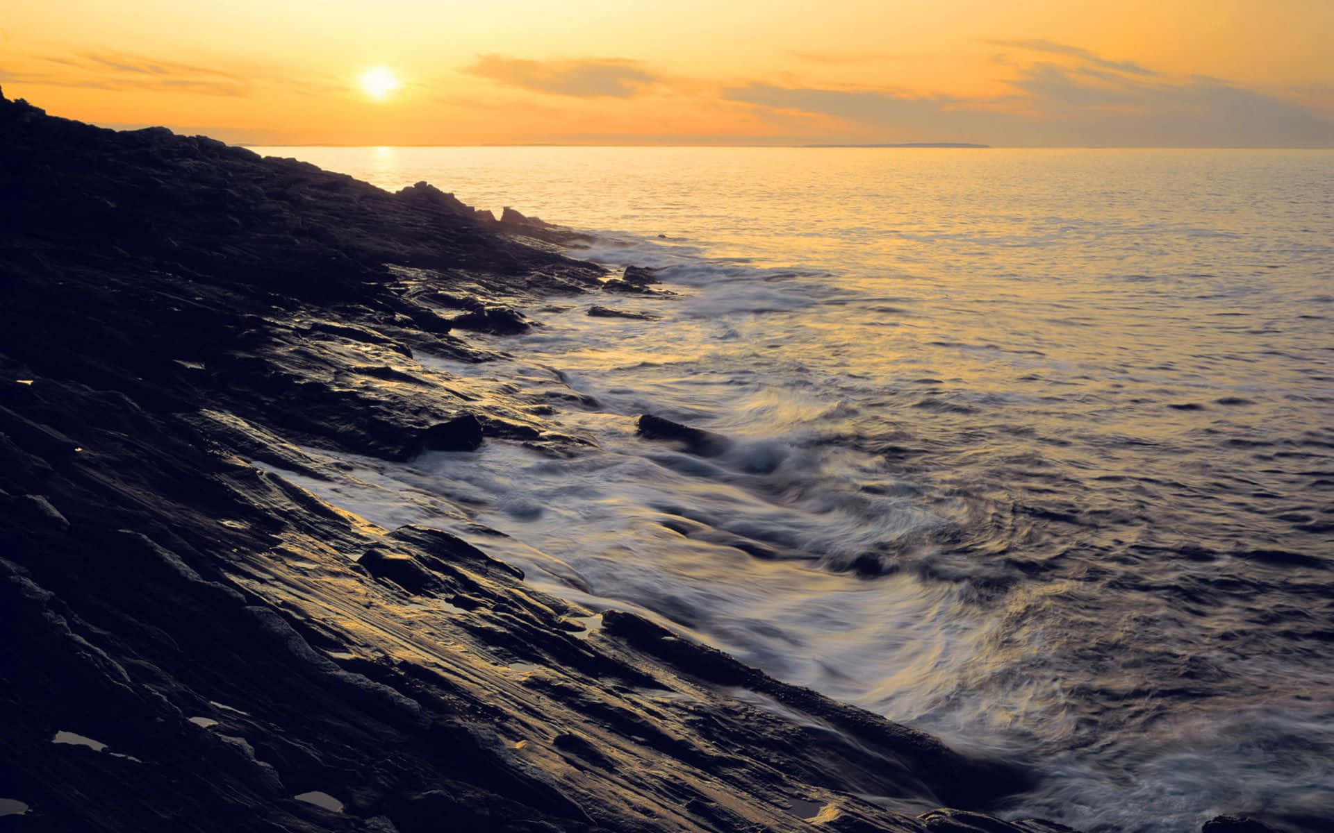 Coastal Sunset 桌布 壁紙 — 金色夕陽將溫暖光線投射在拍打黑暗岩石海岸的波浪上。