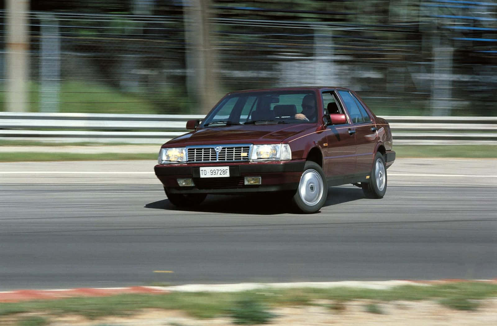  壁紙 — 一輛酒紅色的Lancia Thema轎車在賽道上疾馳，背景呈現動態模糊效果。