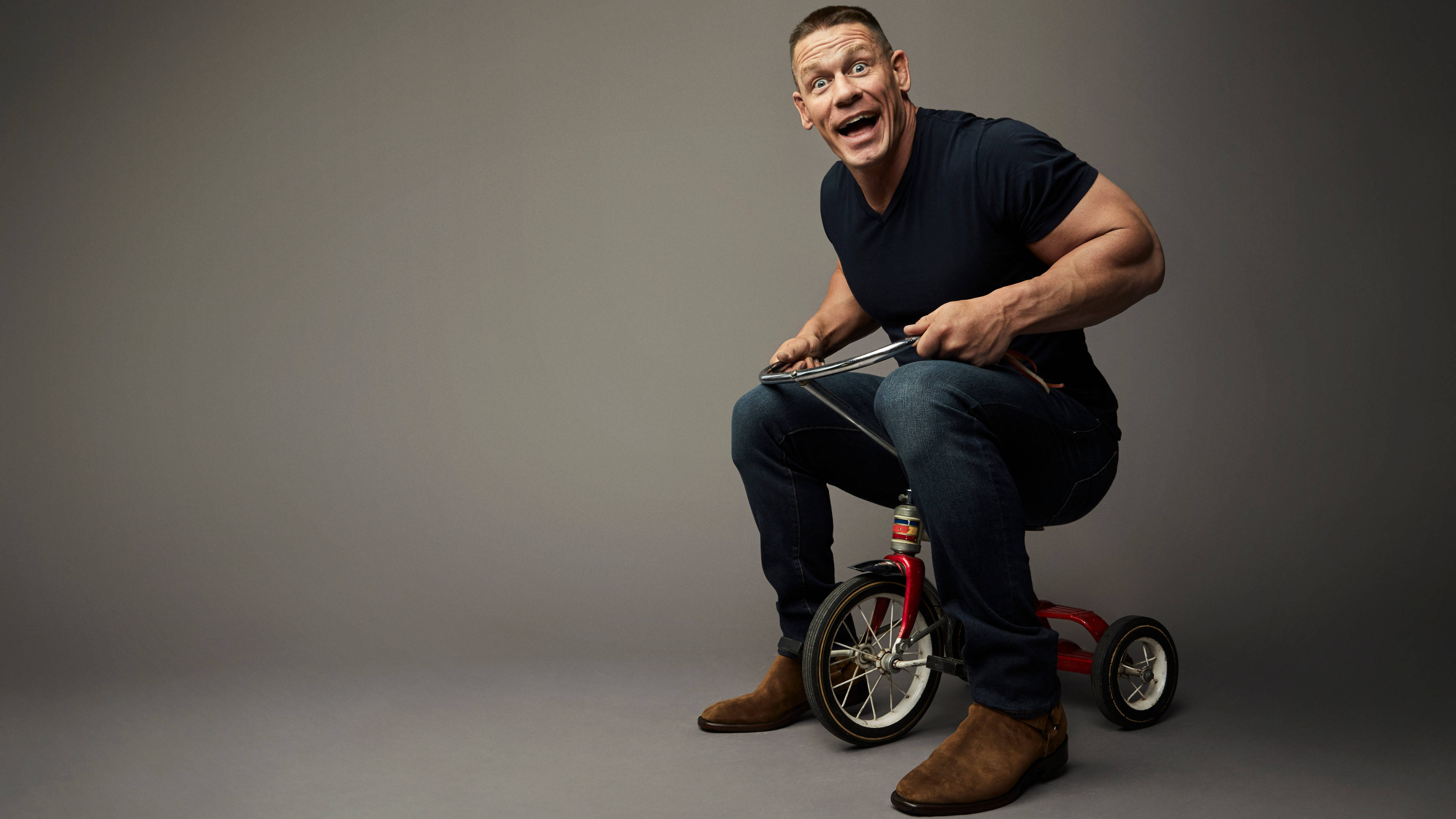 約翰·希南 桌布 壁紙 — John Cena 在純灰色工作室背景前騎紅色三輪車，頑皮地咧嘴笑。