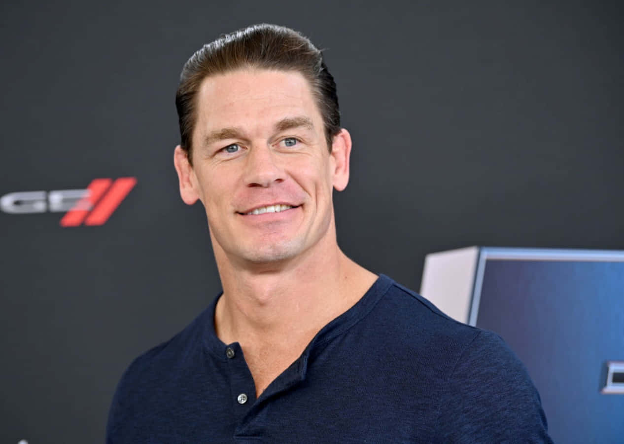 約翰·希南 桌布 壁紙 — John Cena 穿著深藍色亨利衫，在品牌活動背景前溫暖微笑。