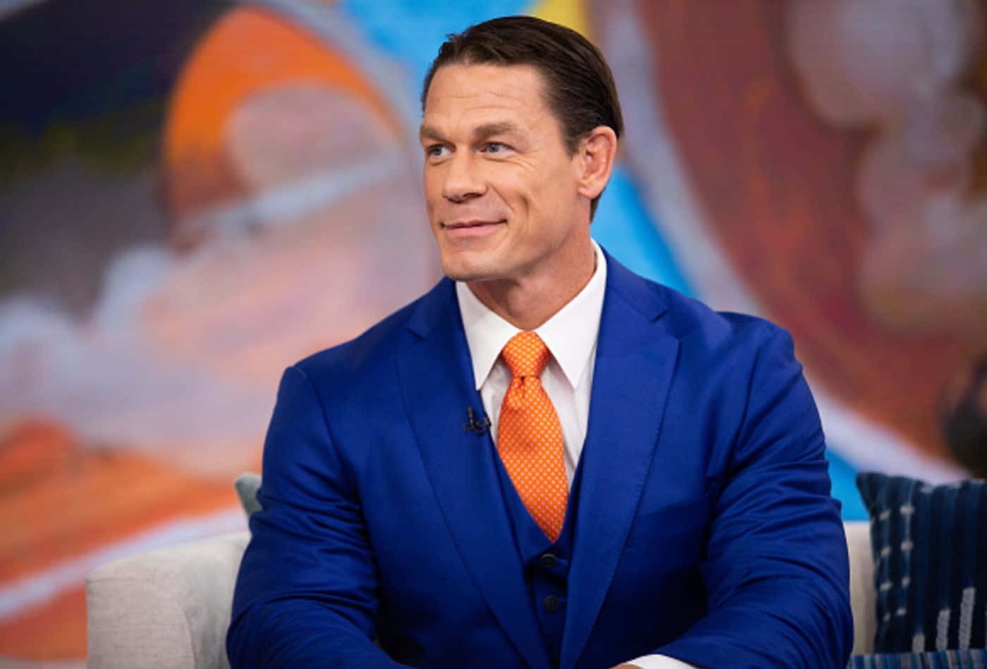 約翰·希南 桌布 壁紙 — John Cena 自信地坐在鮮藍色西裝和橙色領帶中，背景是色彩繽紛的模糊效果。