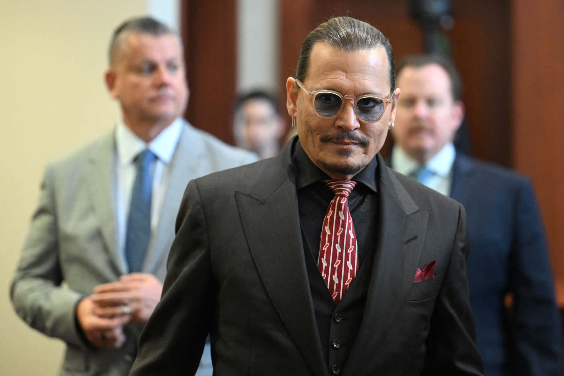 Celebrity Humor Memes 桌布 壁紙 — Johnny Depp 自信地穿著深色西裝和紅色圖案領帶，戴著太陽眼鏡在室內行走，身後有模糊的男人。