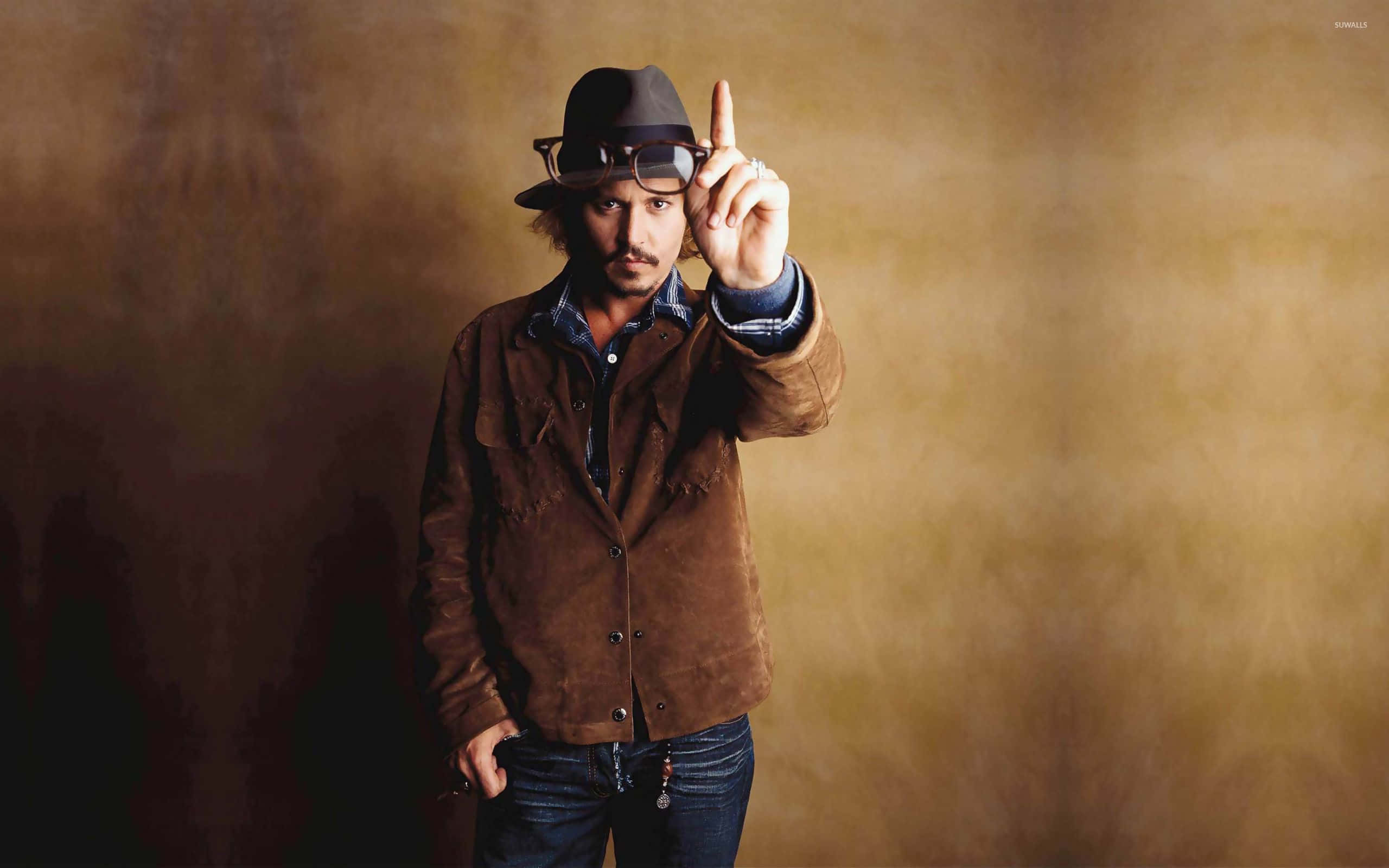 強尼·戴普 桌布 壁紙 — Johnny Depp 身穿棕色麂皮外套和軟呢帽，在溫暖的漸變背景前自信地微笑並向上指。