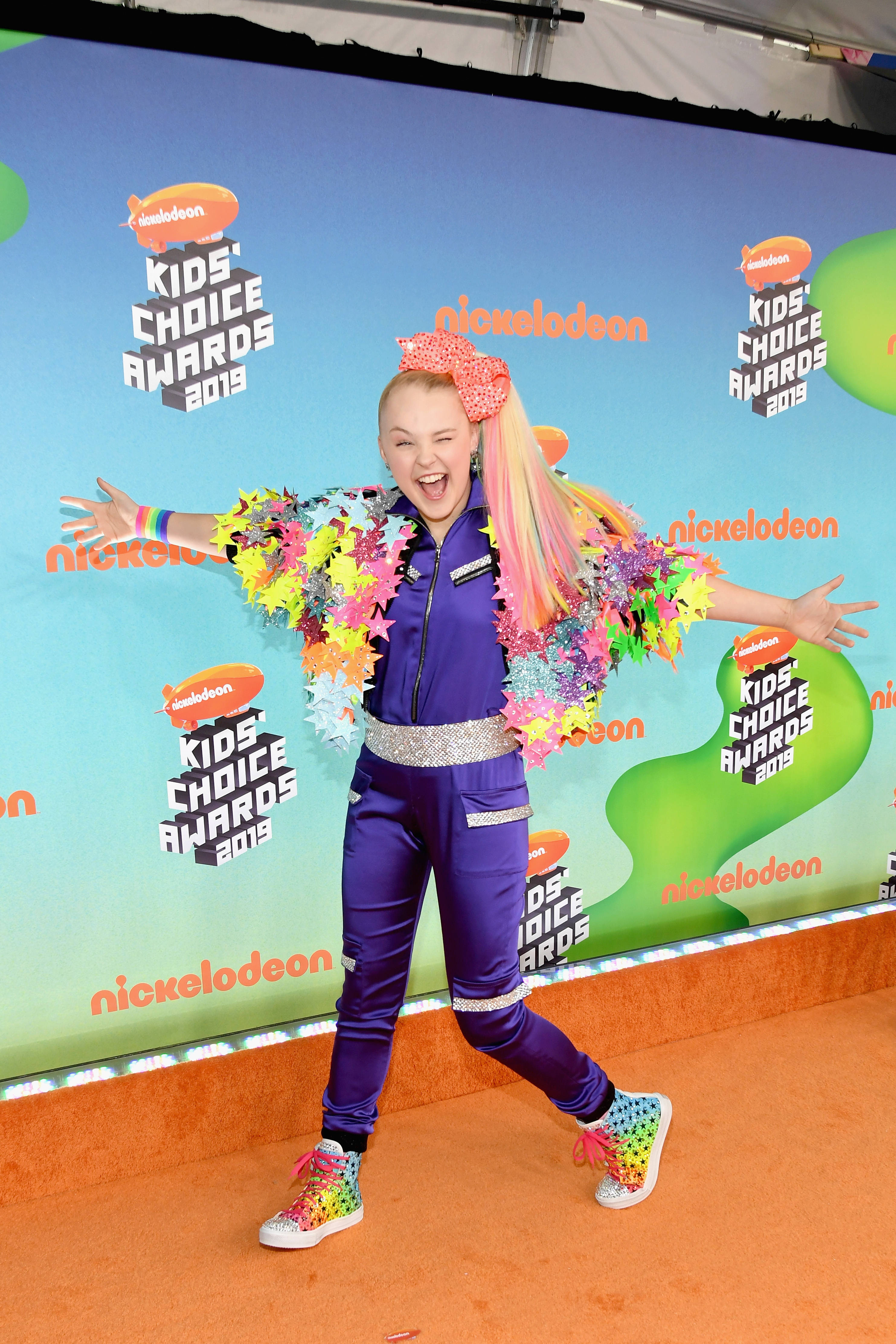 JoJo Siwa 桌布 壁紙 — JoJo Siwa 在2019年兒童選擇獎的橙色地毯上，穿著鮮豔的紫色服裝和彩虹配飾，擺出開心的姿勢。