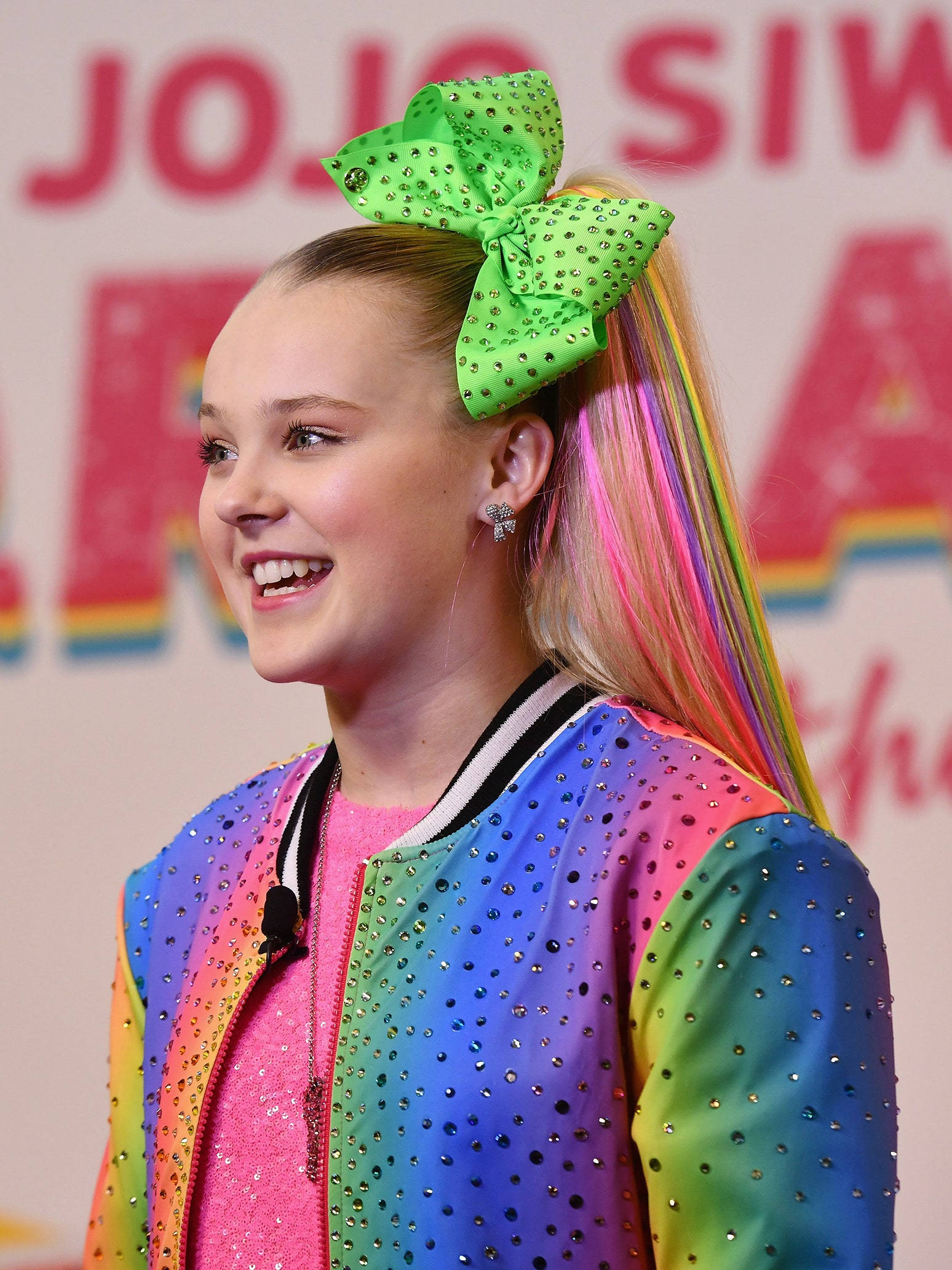 Jojo Siwa 桌布 壁紙 — JoJo Siwa 穿著彩虹亮片外套，色彩繽紛的馬尾上繫著一個巨大的綠色蝴蝶結，露出燦爛的笑容。