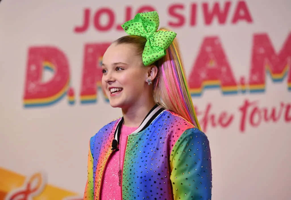 Jojo 桌布 壁紙 — JoJo Siwa 穿著彩虹亮片外套，色彩繽紛的馬尾上繫著一個巨大的綠色圓點蝴蝶結，露出燦爛的笑容。