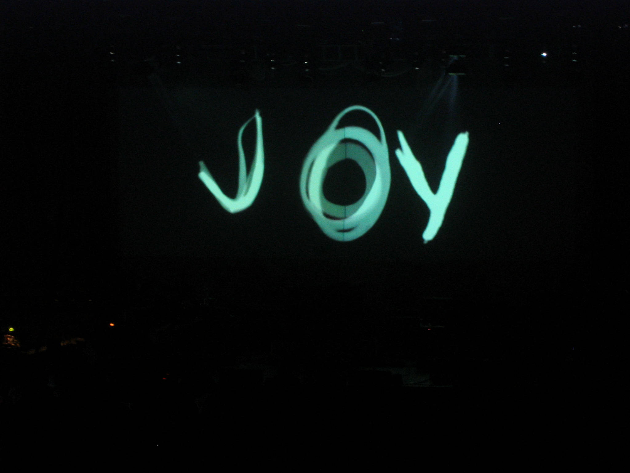 喜悅 桌布 壁紙 — 「JOY」這個詞在漆黑的舞台上散發著充滿活力的藍綠色光芒，在黑暗的音樂會場地被聚光燈照亮。