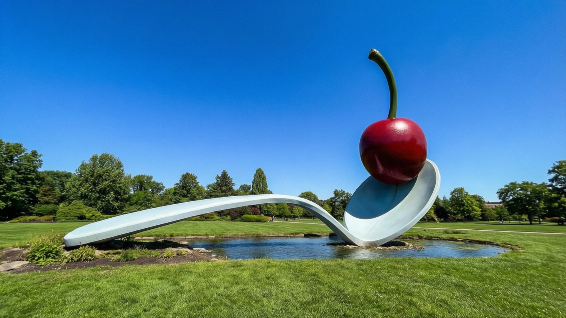 Spoonbridge and Cherry 壁紙 — 一個巨大的銀色湯匙雕塑，上面有一顆光澤的紅色櫻桃平衡在湯匙碗上，位於池塘中，周圍是綠草和樹木，在清澈的藍天下。