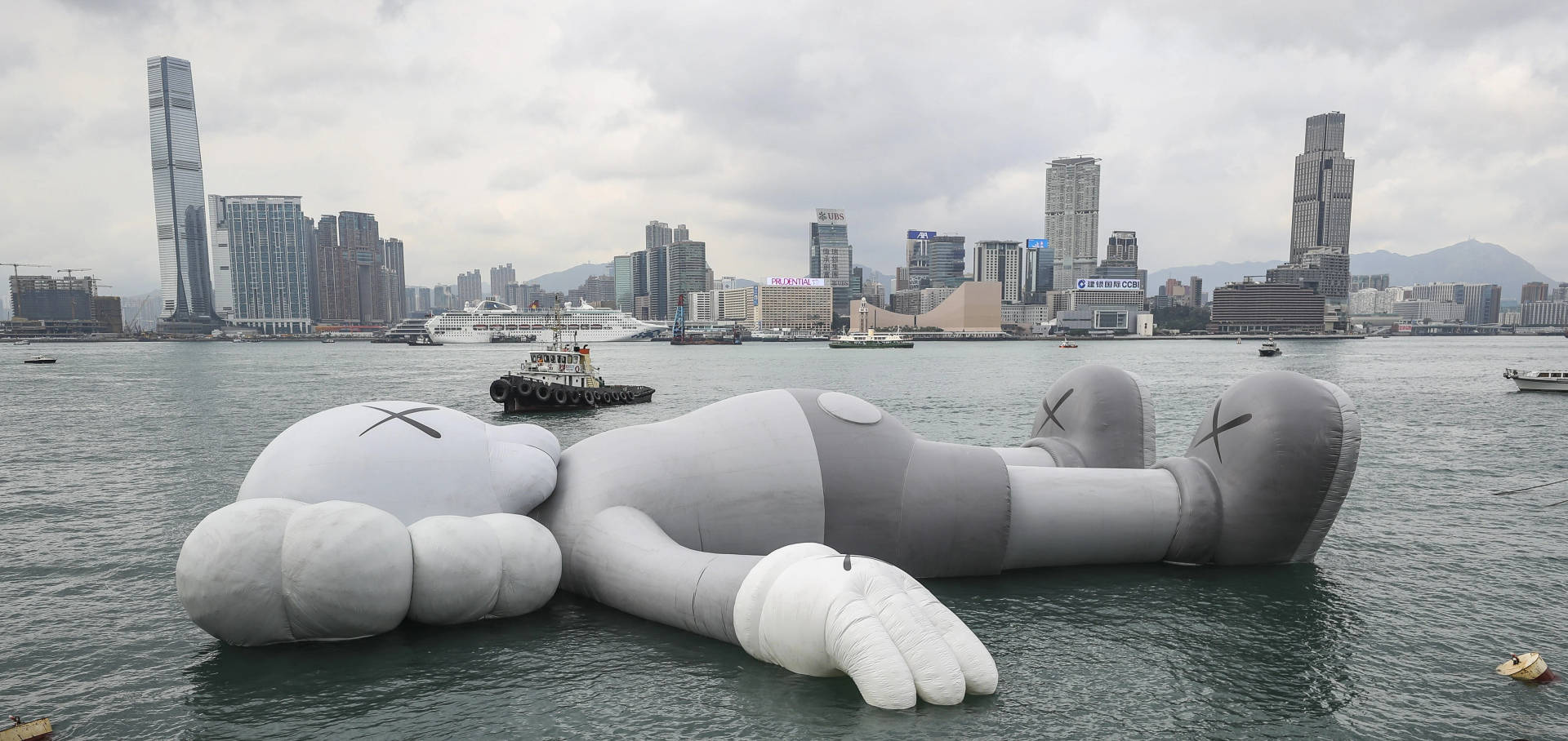 大型灰色KAWS雕塑漂浮在港口水中，背景為城市天際線。 — Kaws 壁紙
