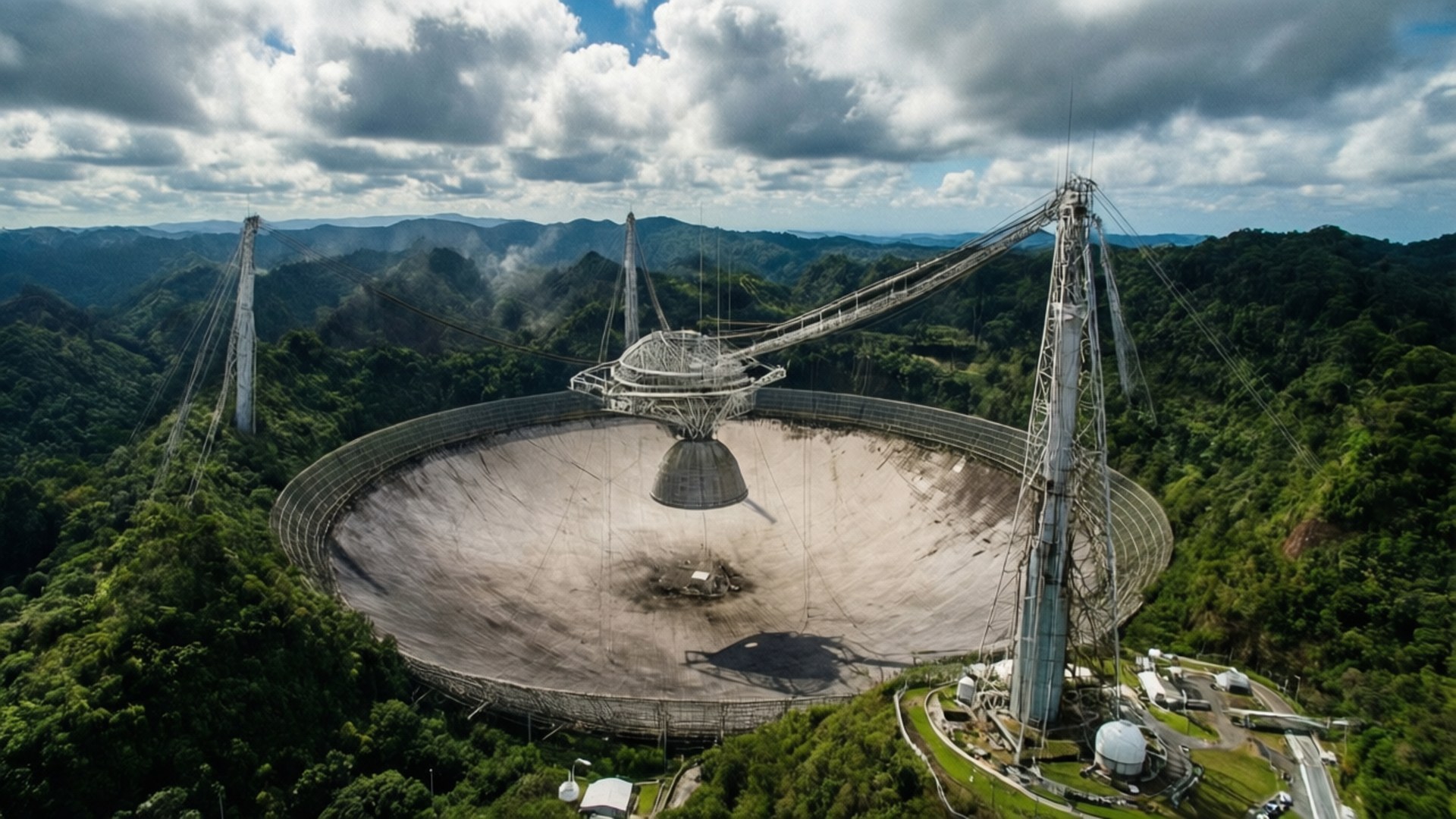 Arecibo Observatory Aerial 壁紙 — 一個巨大的無線電望遠鏡碟形天線，坐落在鬱鬱蔥蔥的綠色山谷中，上方是戲劇性的多雲天空。