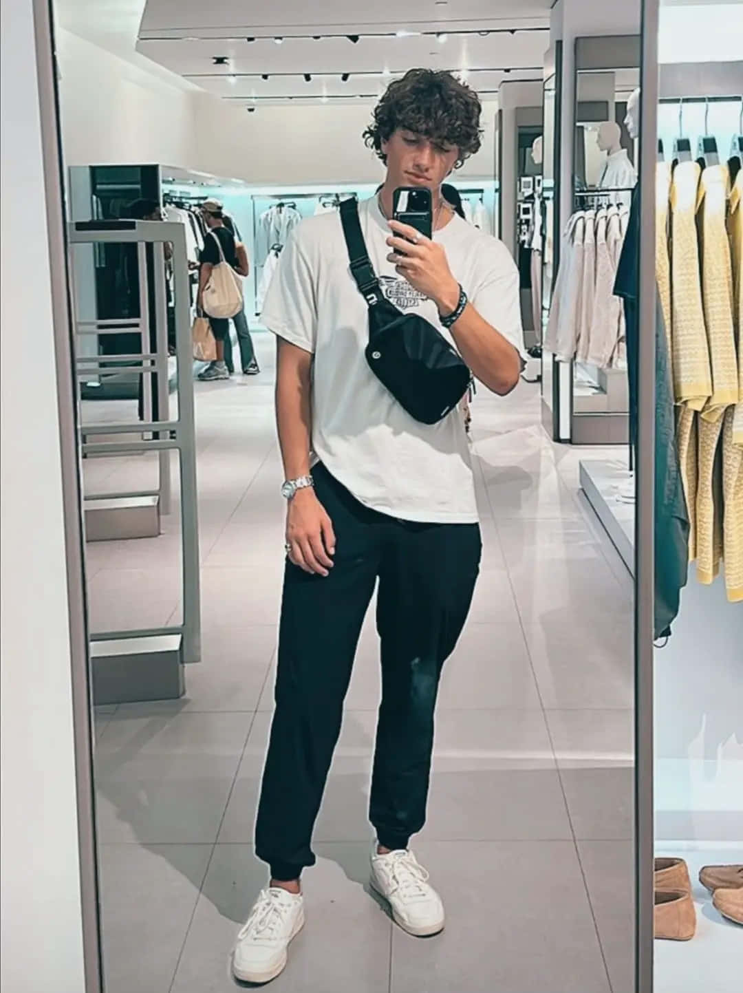  壁紙 — 一位捲髮年輕男子在明亮的服裝店裡，穿著白色T恤、黑色褲子和白色運動鞋，搭配黑色腰包，對著鏡子自拍。