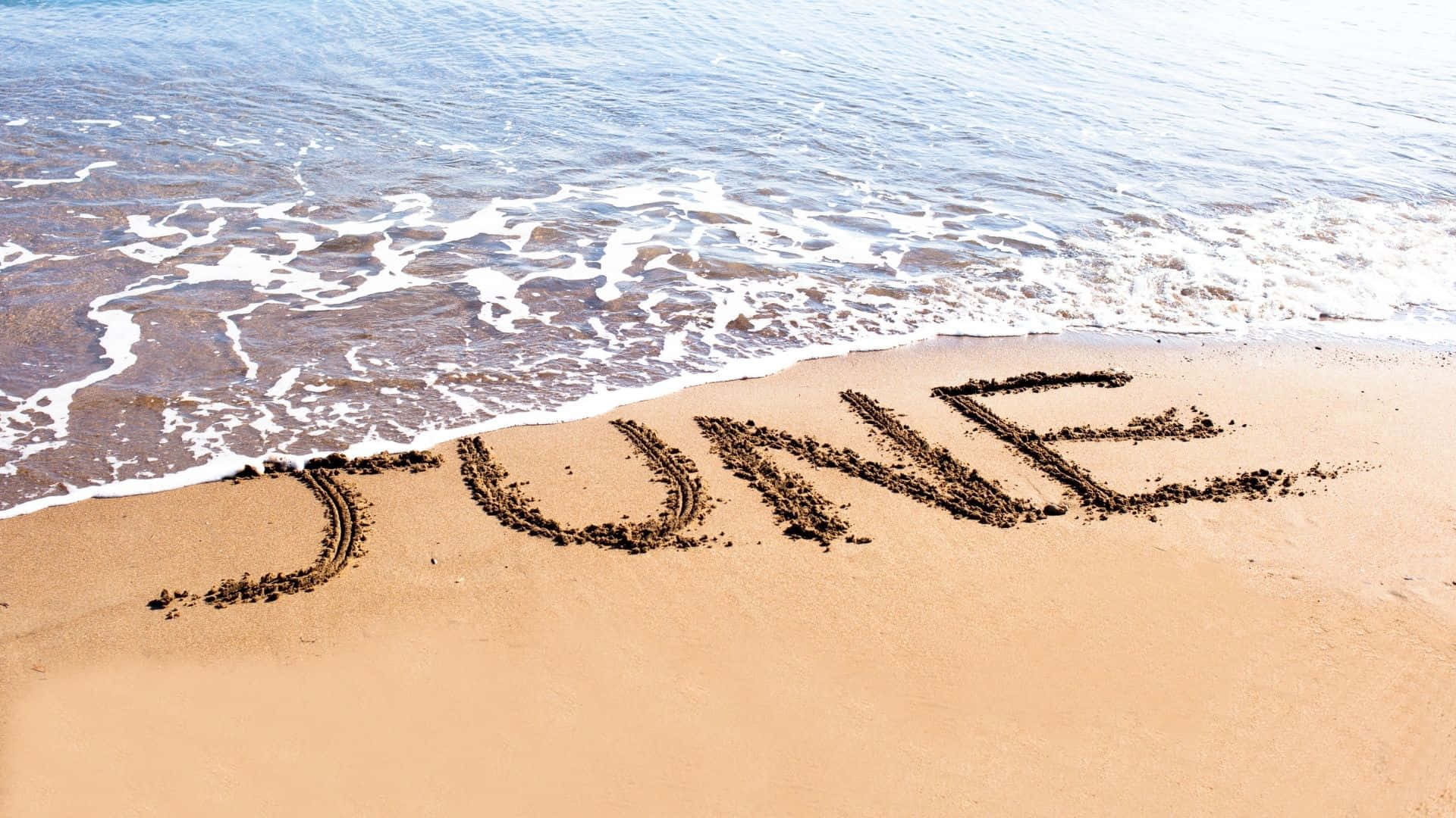June 桌布 壁紙 — 「JUNE」這個字寫在深色沙灘的水邊，溫柔的海浪輕輕拍打字母。