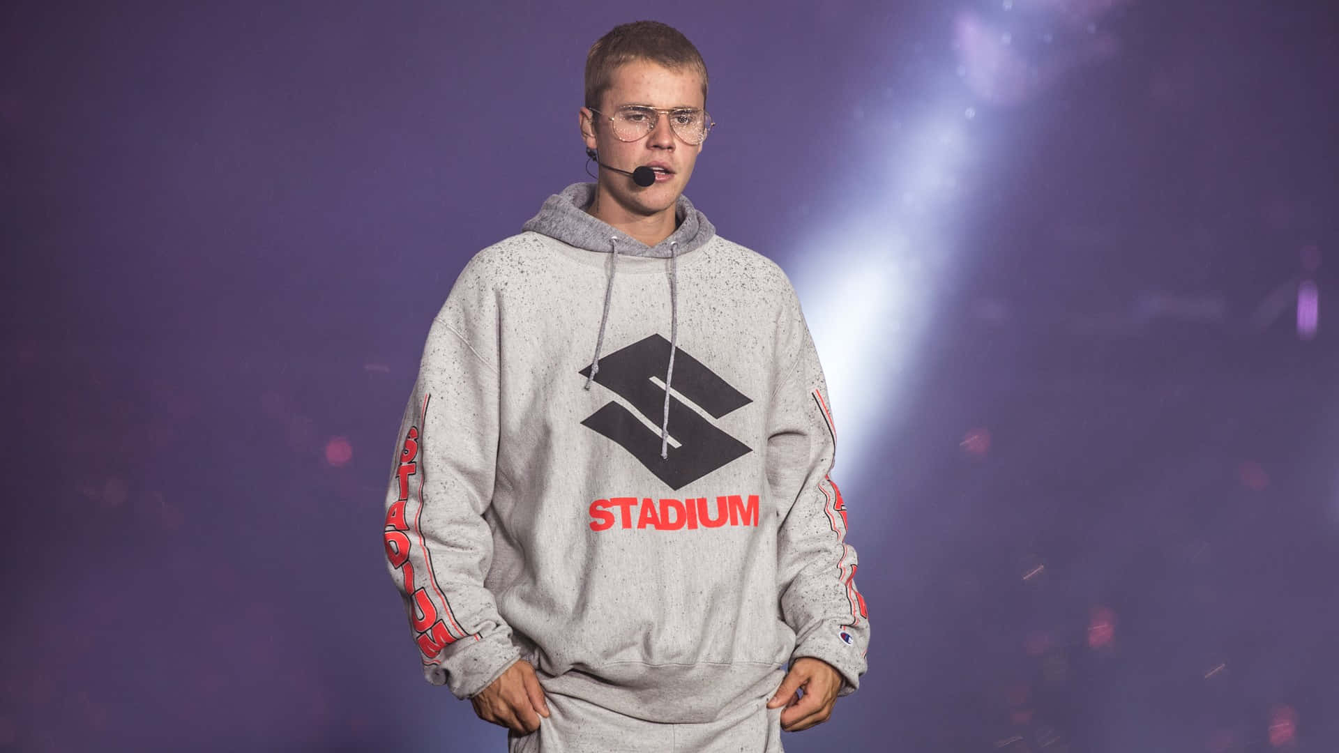 Justin Bieber 2021 桌布 壁紙 — Justin Bieber 在紫色舞台燈光下，身穿灰色連帽衫，上面有紅色 'STADIUM' 字樣，站在舞台上。