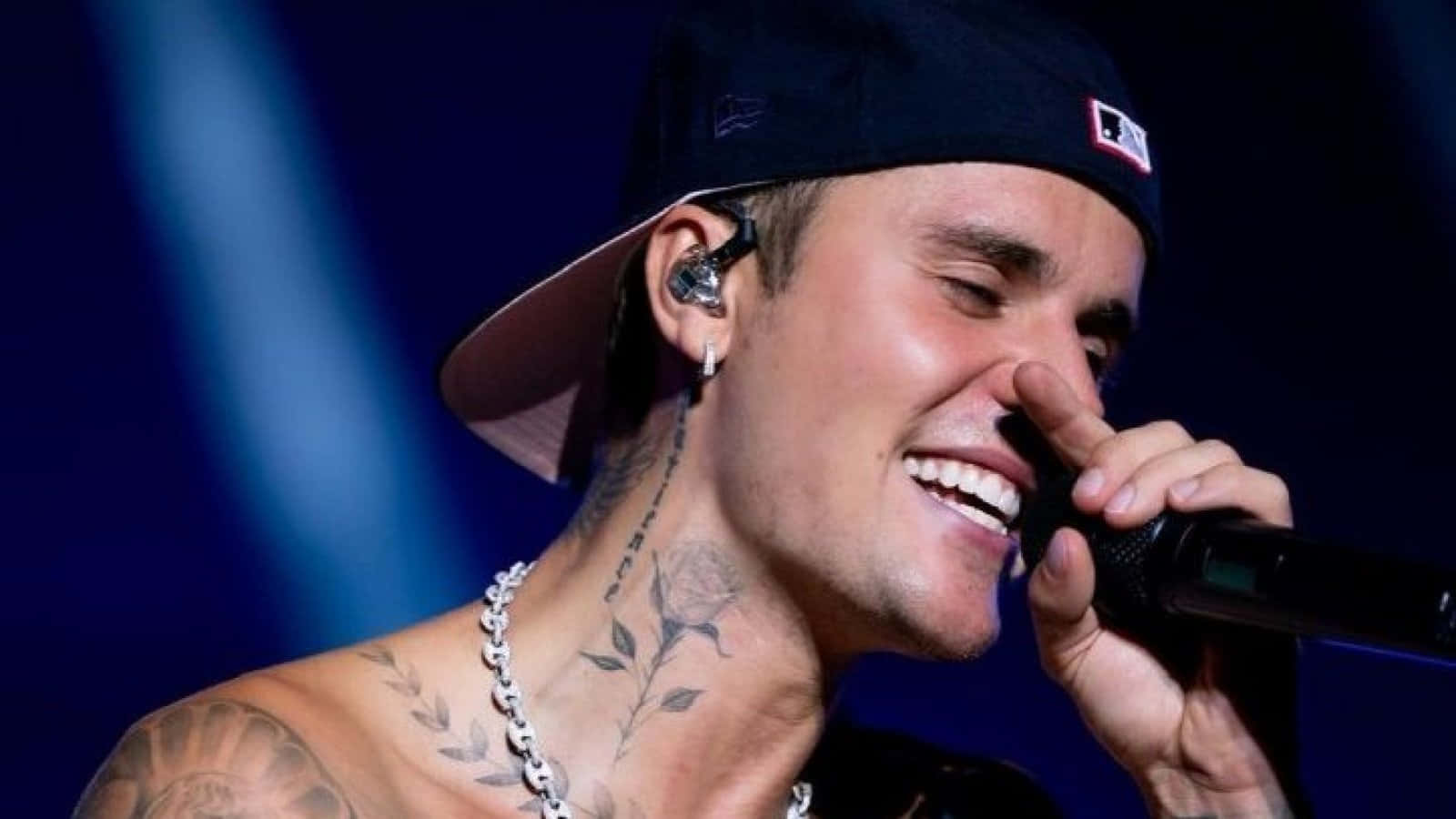 Tattoo 桌布 壁紙 — Justin Bieber 在藍色燈光的舞台上對著麥克風快樂地唱歌大笑，可見刺青和帽子。