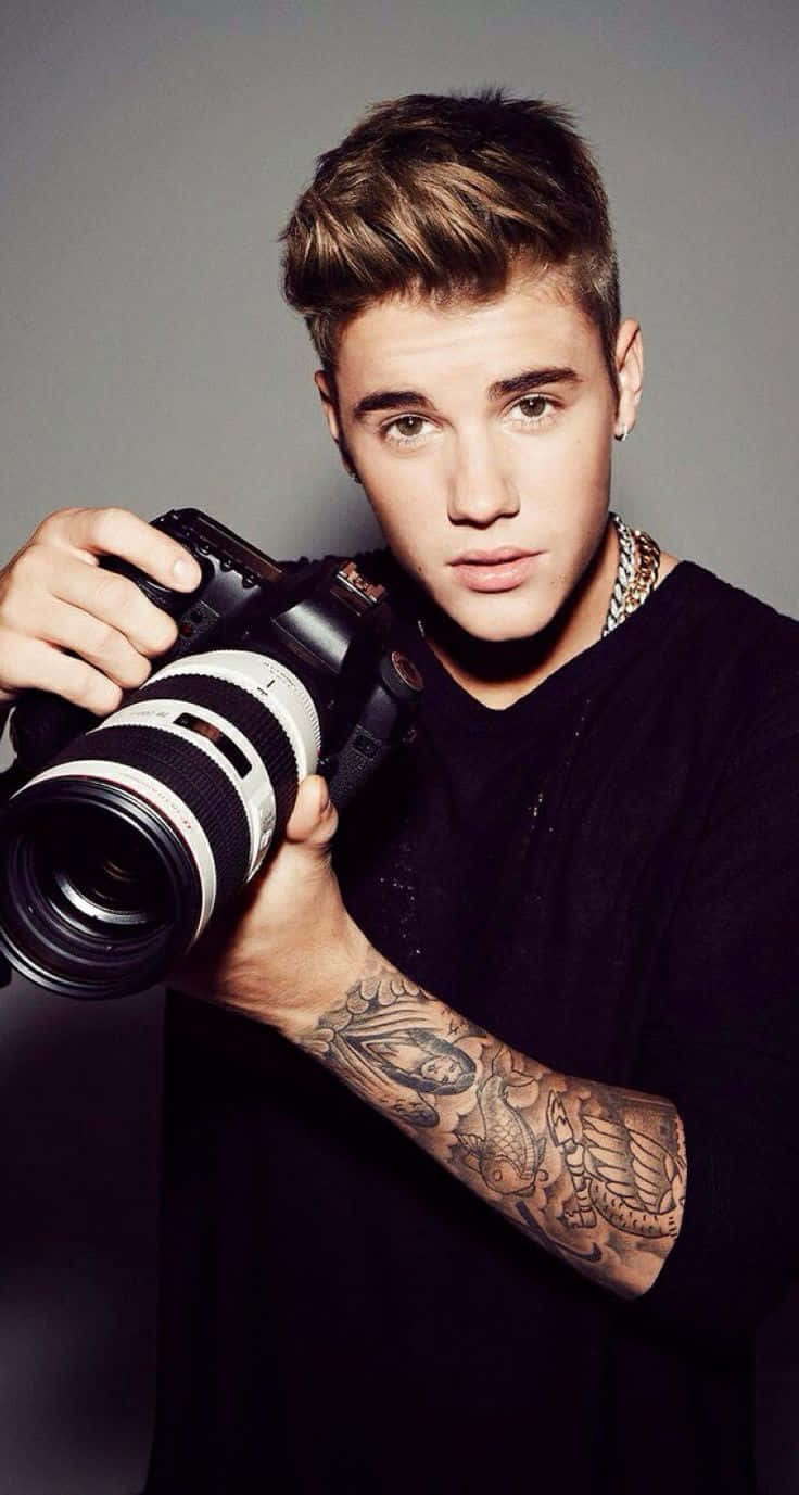 小賈斯汀 桌布 壁紙 — Justin Bieber 手持一部配備大鏡頭的 DSLR 相機，在純灰色背景前直視觀眾。