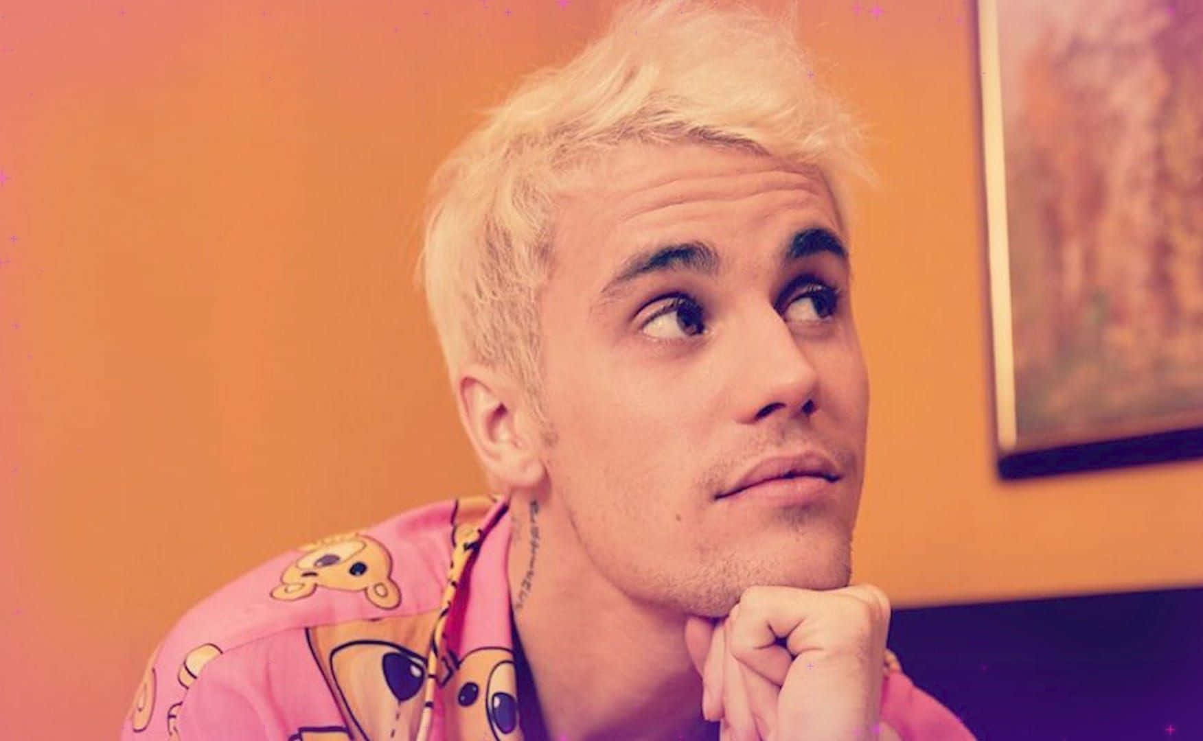 小賈斯汀 桌布 壁紙 — Justin Bieber 留著鉑金色頭髮，身穿粉紅色卡通襯衫，將下巴擱在手上，靠著溫暖的橙色牆壁。