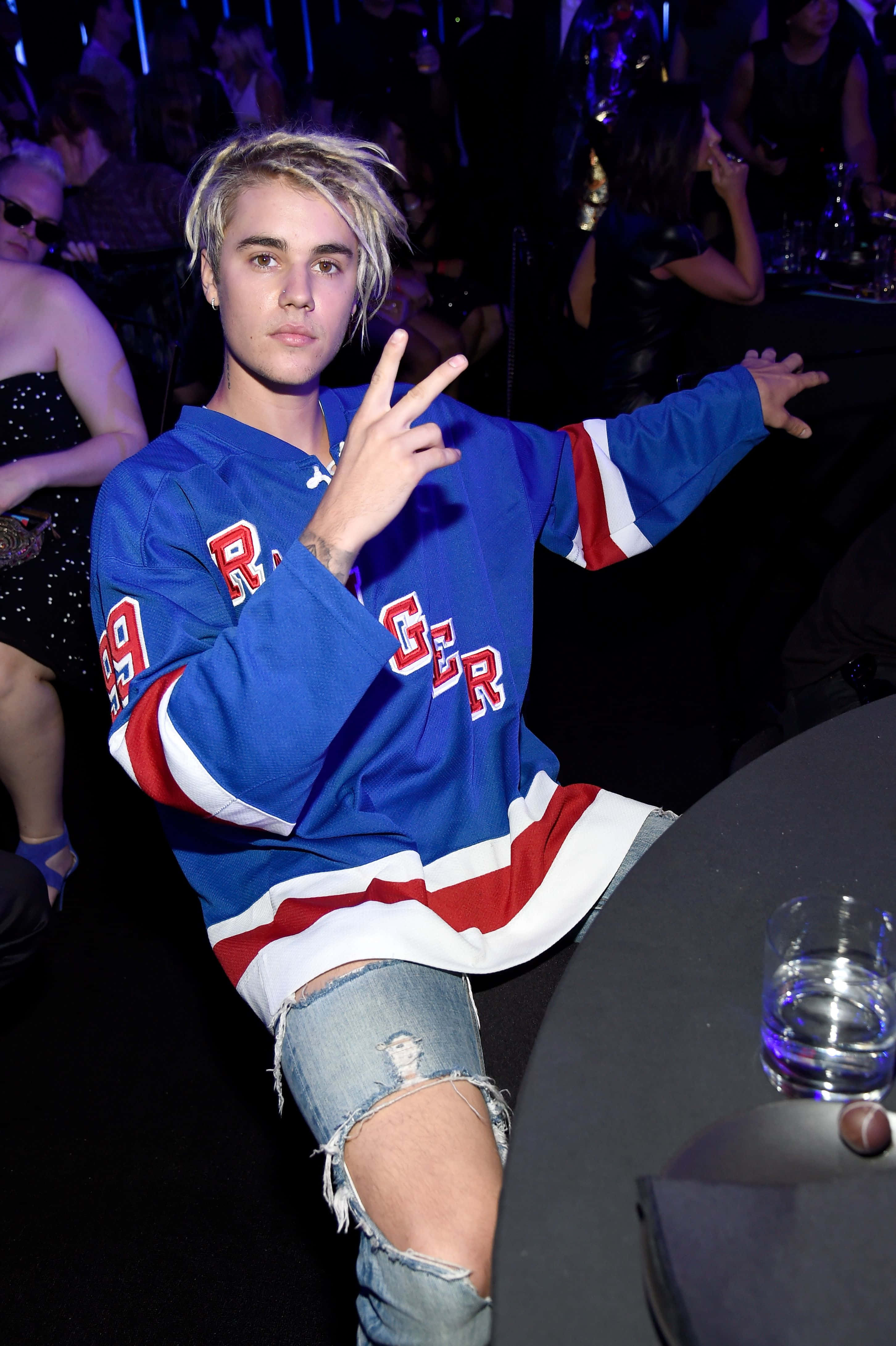 Justin Bieber 2021 桌布 壁紙 — Justin Bieber 自信地擺姿勢，身穿藍、紅、白色冰球球衣，在燈光昏暗的派對中，背後有閃爍的燈光和人群。