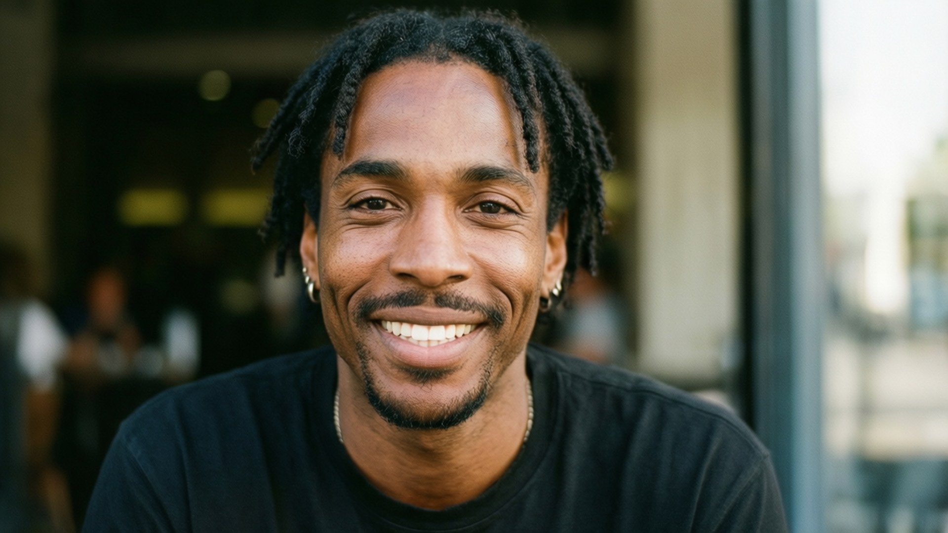 Friendly Smile Portrait 壁紙 — 一個開朗的年輕男子留著辮子，坐在玻璃門附近的戶外，對著鏡頭溫暖地微笑。
