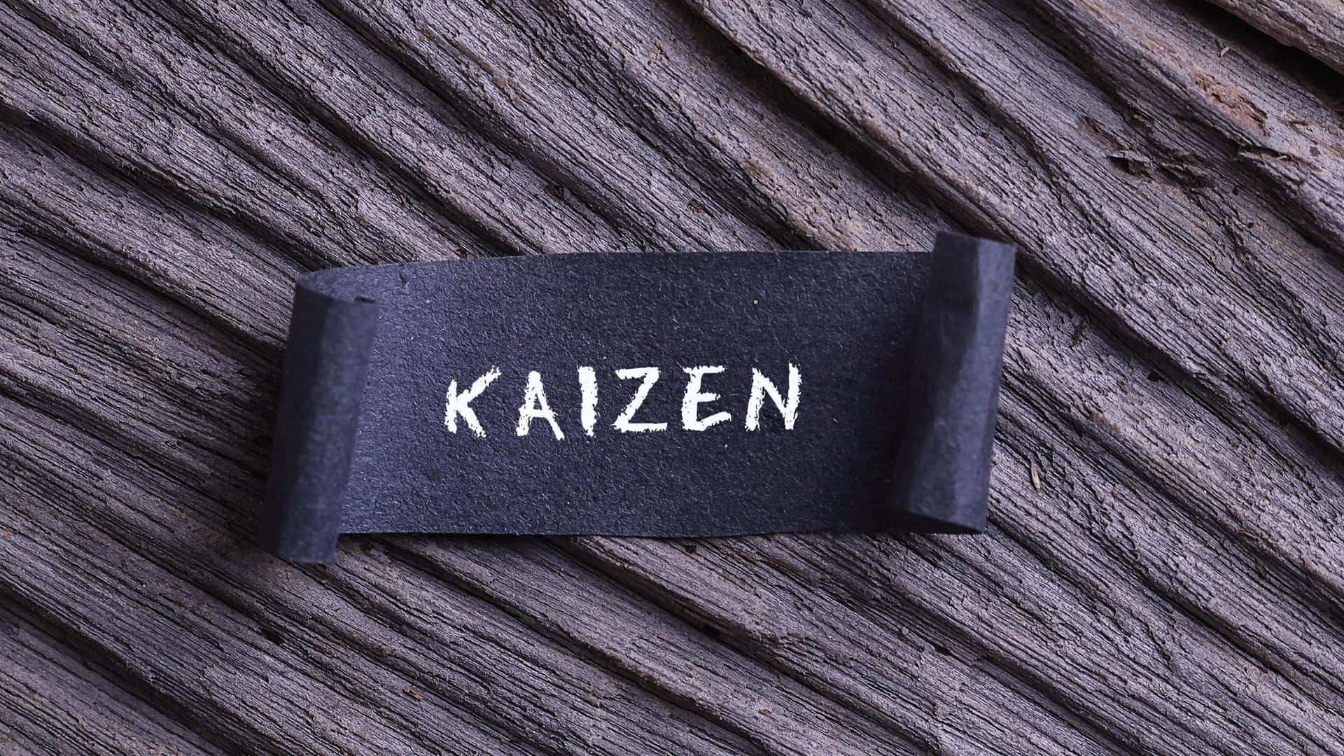  壁紙 — 「KAIZEN」這個字用白色粉筆寫在一條黑色黑板條上，而黑板條則放在經風化的灰色木頭上。