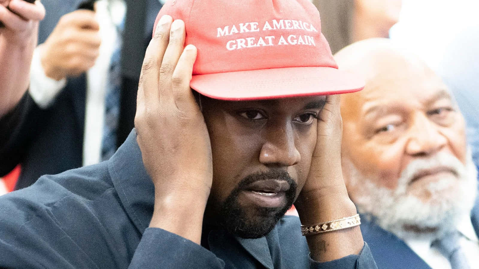 Kanye 桌布 壁紙 — Kanye West 在公開活動中戴著紅色 'Make America Great Again' 帽子，同時痛苦地抱著頭。