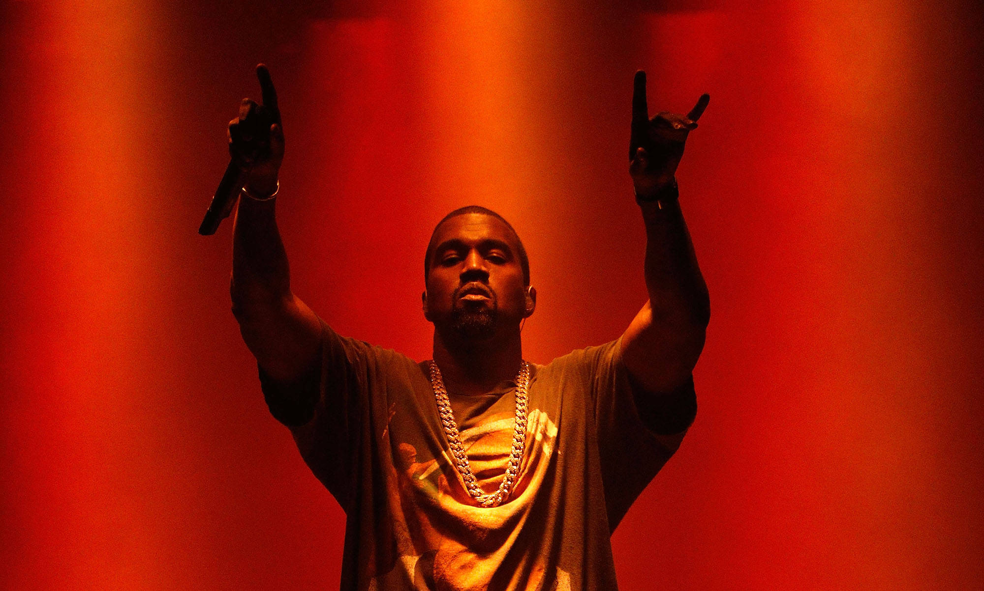 Kanye 桌布 壁紙 — Kanye West 在強烈紅燈下舉起雙臂，手持麥克風在舞台上表演。