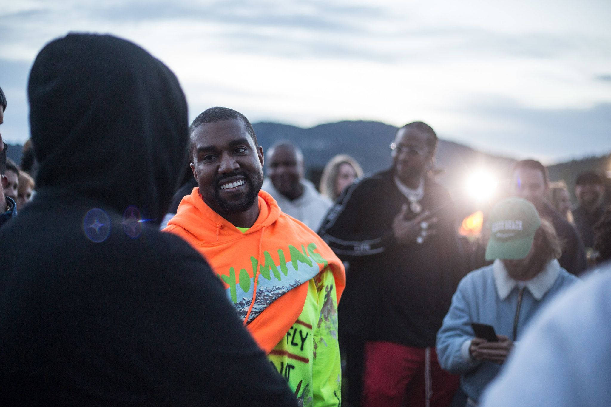 Kanye West Ye 桌布 壁紙 — Kanye West 在戶外聚會中，身穿亮橙色和螢光綠色連帽衫，背景是山脈，笑容燦爛。