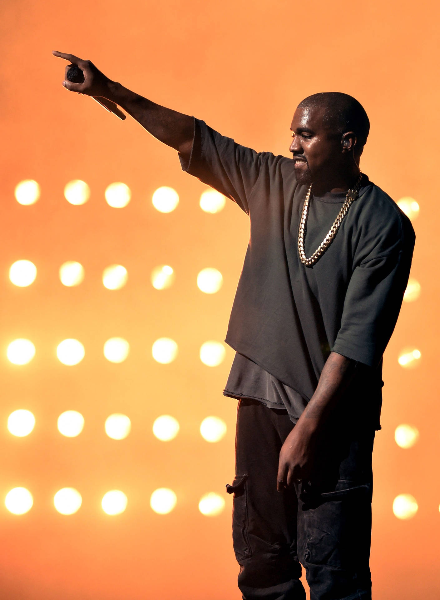  壁紙 — Kanye West 在橙色背景前，在溫暖的燈光下自信地指向舞台。