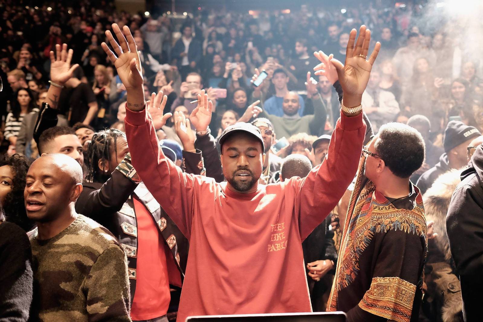 說唱歌手 桌布 壁紙 — Kanye West 在演唱會上，在明亮的舞台燈光下，舉起雙手，身穿紅色運動衫，周圍是歡呼的人群。