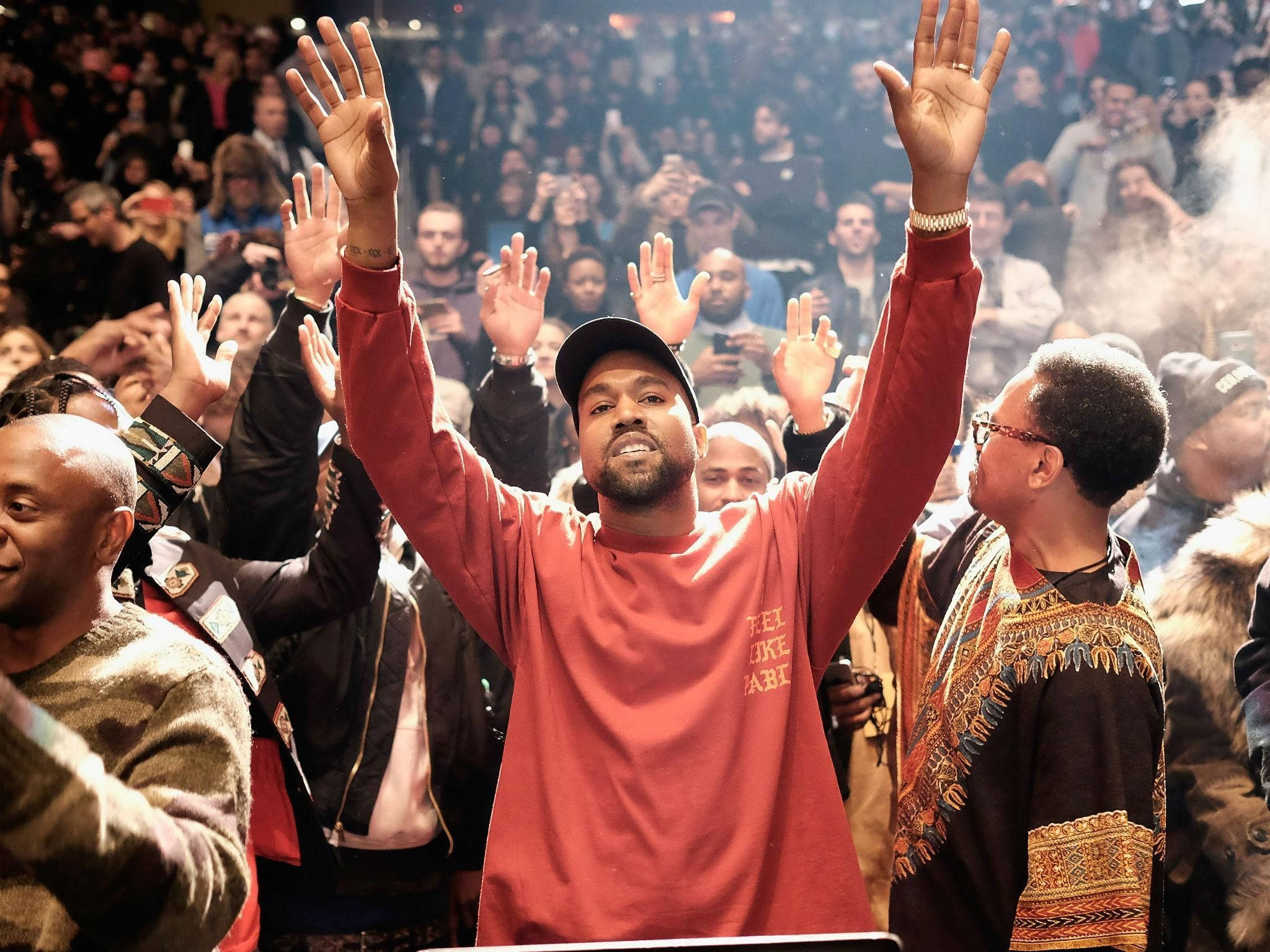 Kanye West 桌布 壁紙 — Kanye West 在演唱會中於煙霧和閃爍燈光下在人群中高舉雙手。