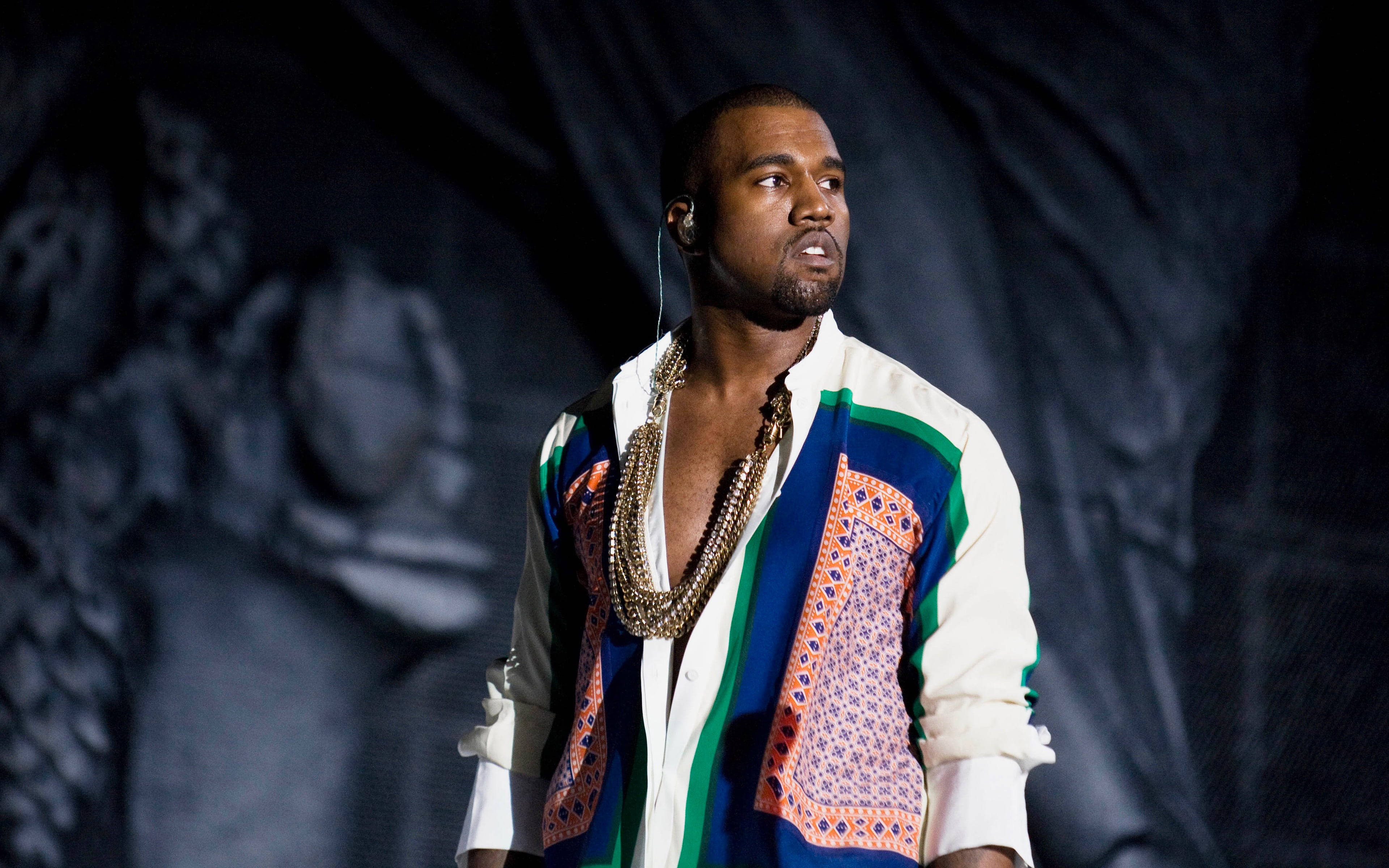 Kanye West Ye 桌布 壁紙 — Kanye West 在舞台上表演，穿著色彩繽紛的圖案背心和層疊項鍊，在戲劇性的舞台燈光下。
