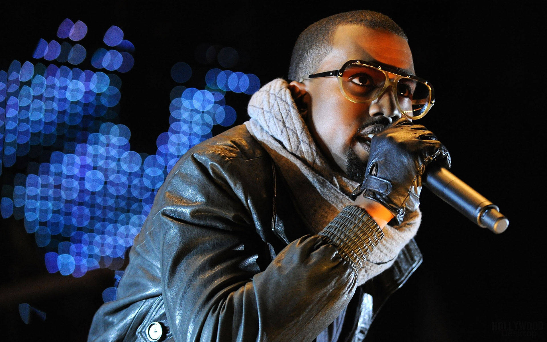Kanye 桌布 壁紙 — Kanye West 在舞台上表演，戴著眼鏡、皮夾克和手套，手持麥克風，背景是藍色散景燈光。