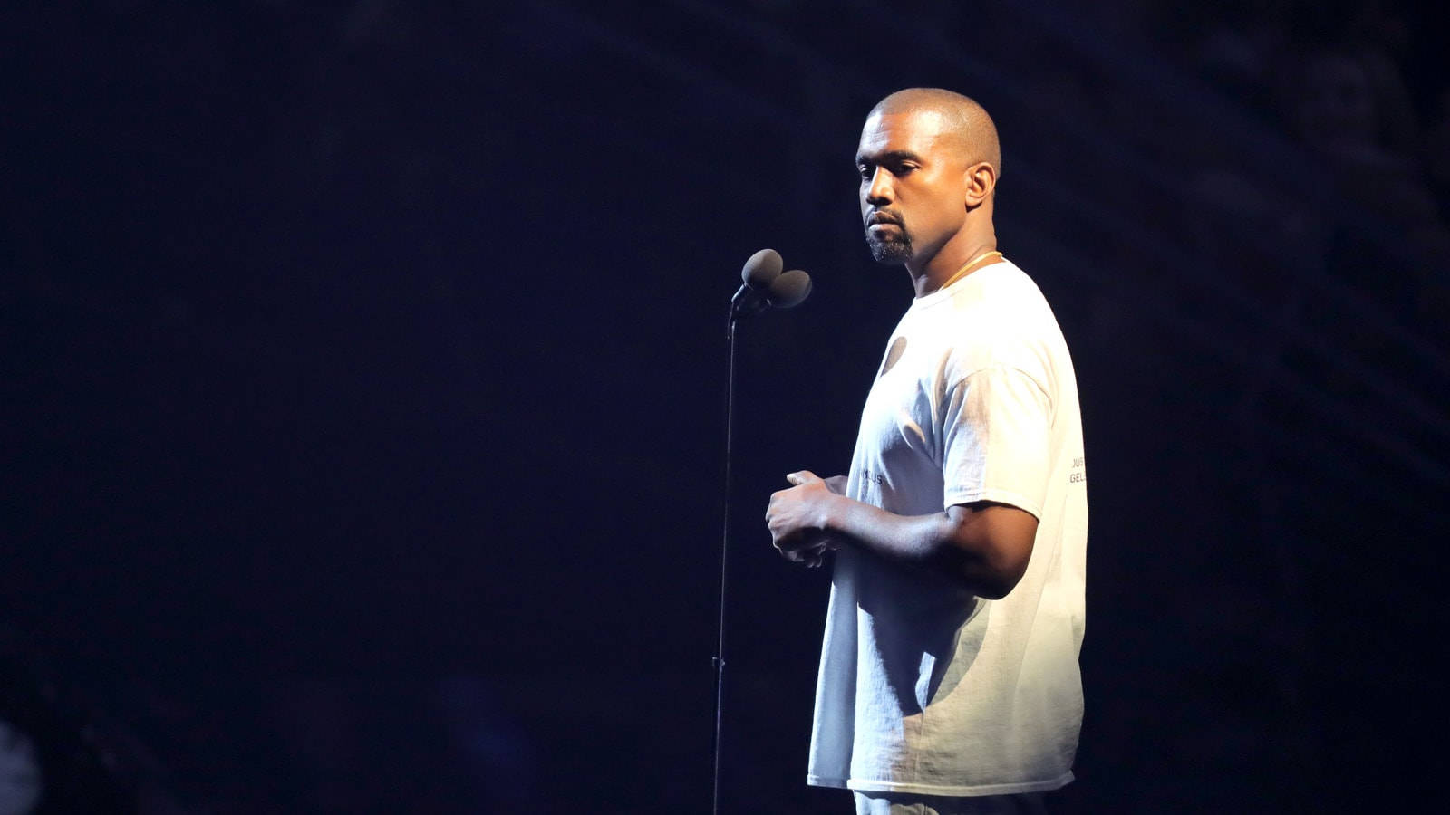 Kanye 桌布 壁紙 — Kanye West 在黑暗舞台上以側面站立，被聚光燈照亮，穿著白色T恤，靠近麥克風。