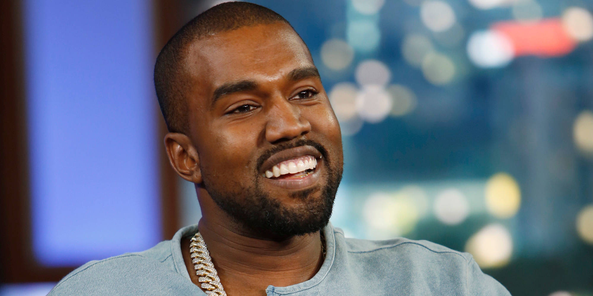Kanye 桌布 壁紙 — Kanye West 戴著鏈條項鍊，在模糊的城市景觀背景下燦爛地微笑。