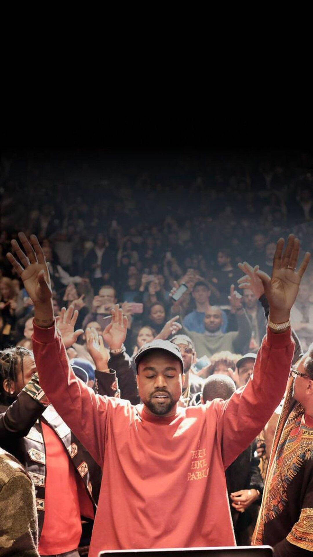 kanye iPhone 桌布 壁紙 — Kanye West 穿著紅色衛衣，在明亮的舞台燈光下向歡呼的人群舉起雙手。