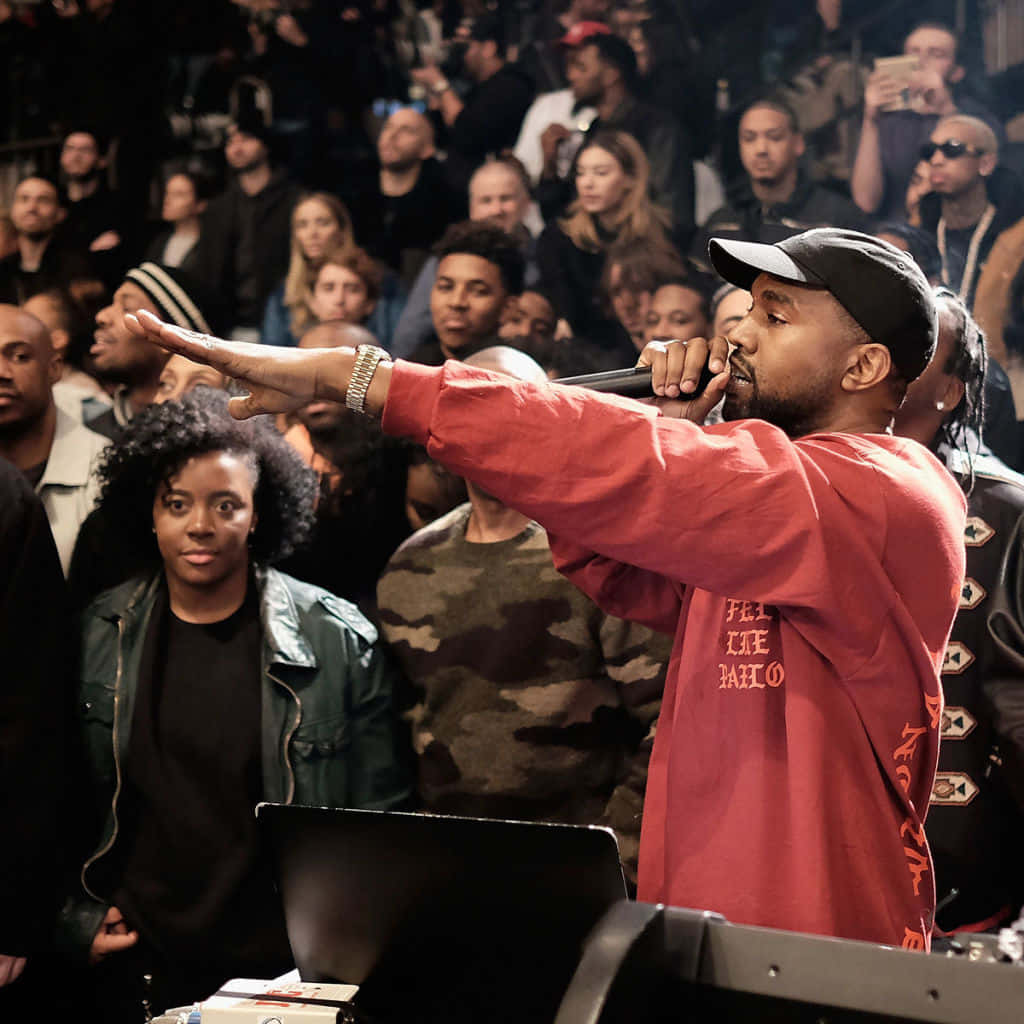 The Life Of Pablo 桌布 壁紙 — Kanye West 穿著紅色襯衫和黑色帽子，在昏暗的場地中熱情地對著麥克風表演，同時指向人群。