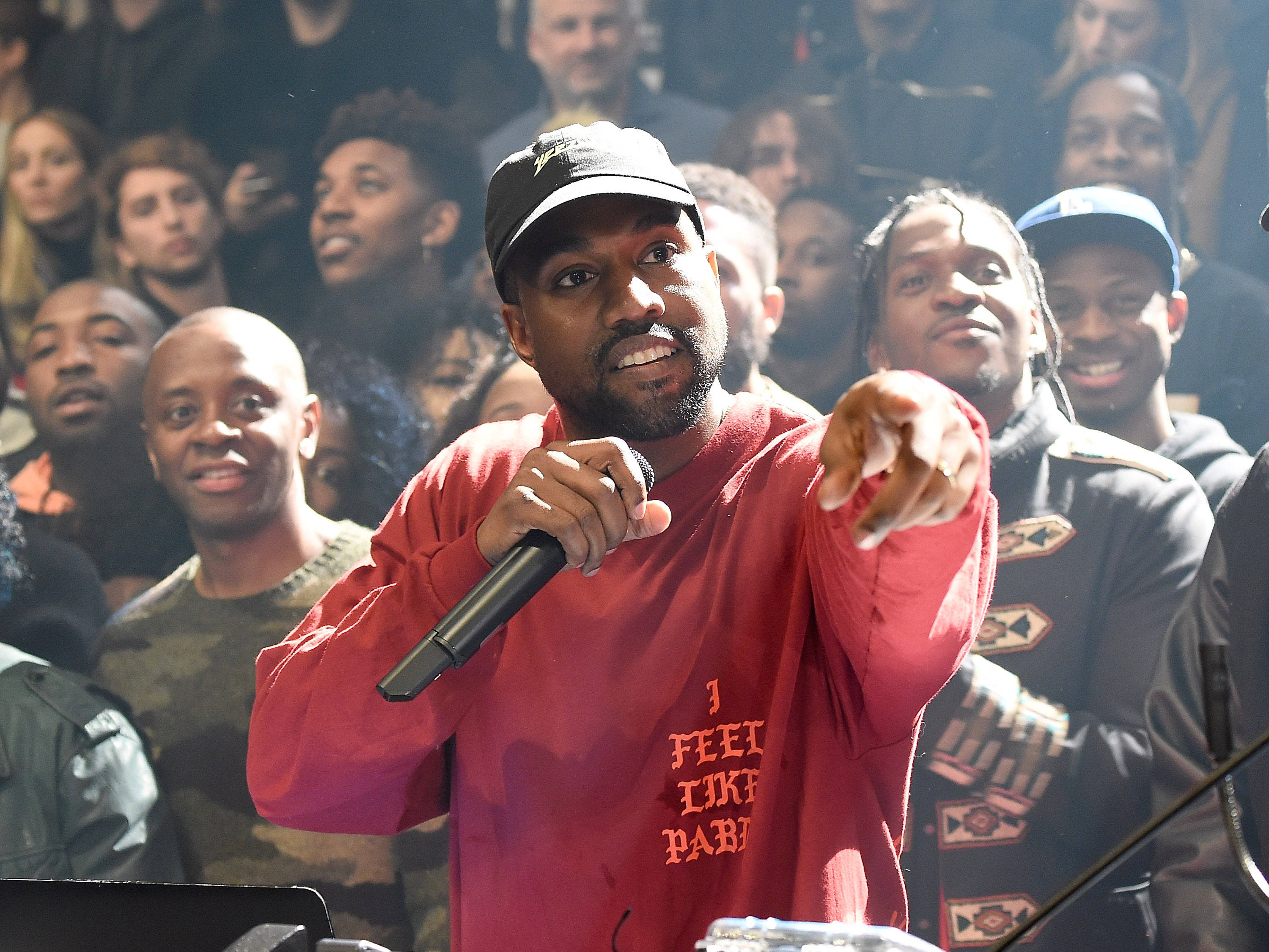 說唱歌手 桌布 壁紙 — Kanye West 穿著紅色連帽衫和帽子，手持麥克風，在微笑的人群中向前指著，在音樂會上。