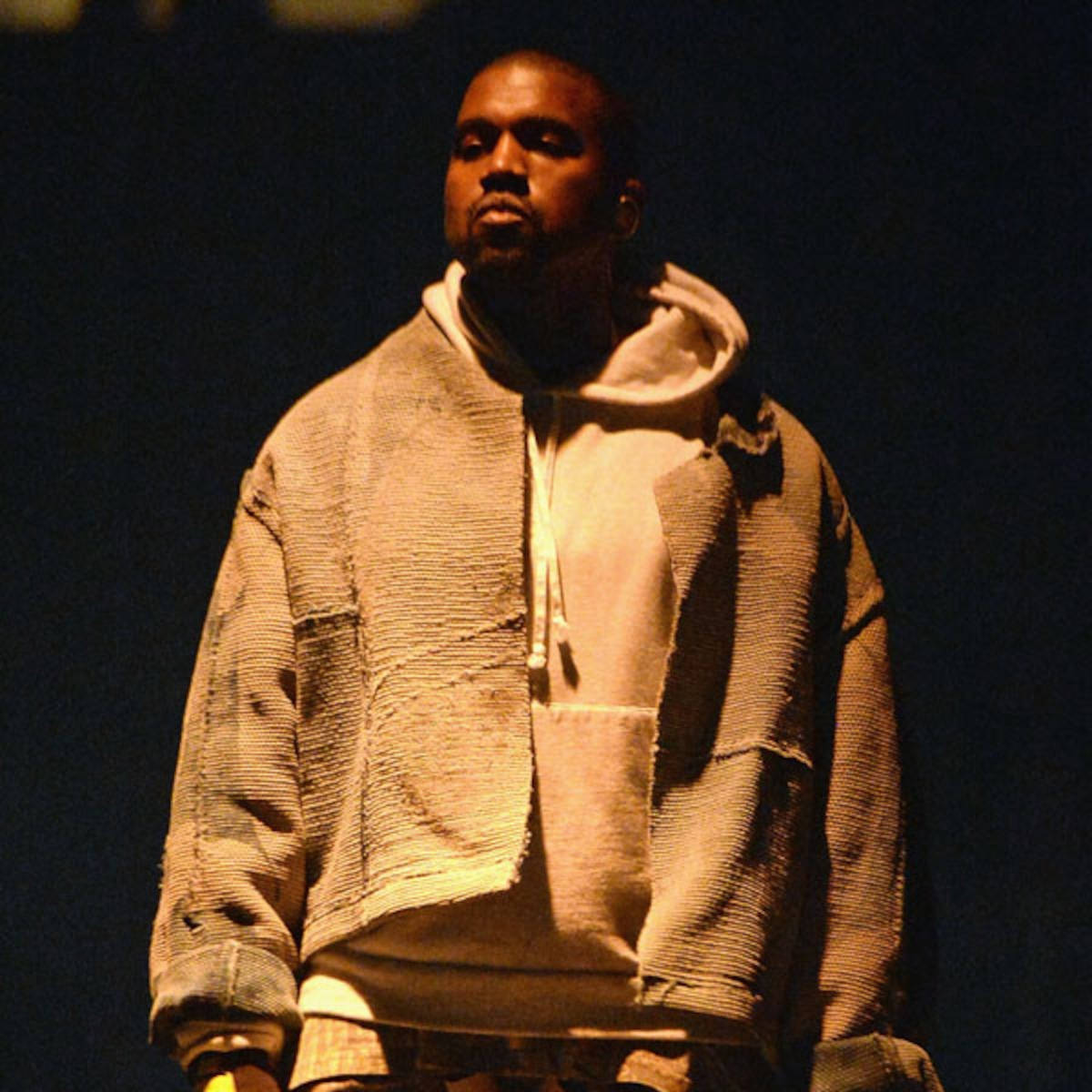 說唱歌手 桌布 壁紙 — Kanye West 站在昏暗的舞台燈光下，穿著一件帶有白色內襯的質感米色連帽衫。
