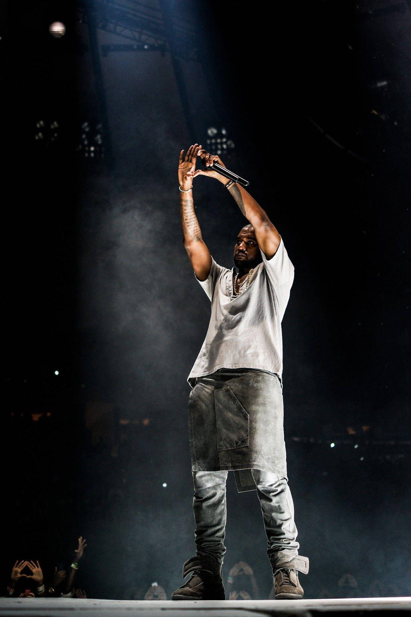 說唱歌手 桌布 壁紙 — Kanye West 站在舞台上，雙臂高舉，握著麥克風，在戲劇性的聚光燈下，煙霧在他周圍旋轉。