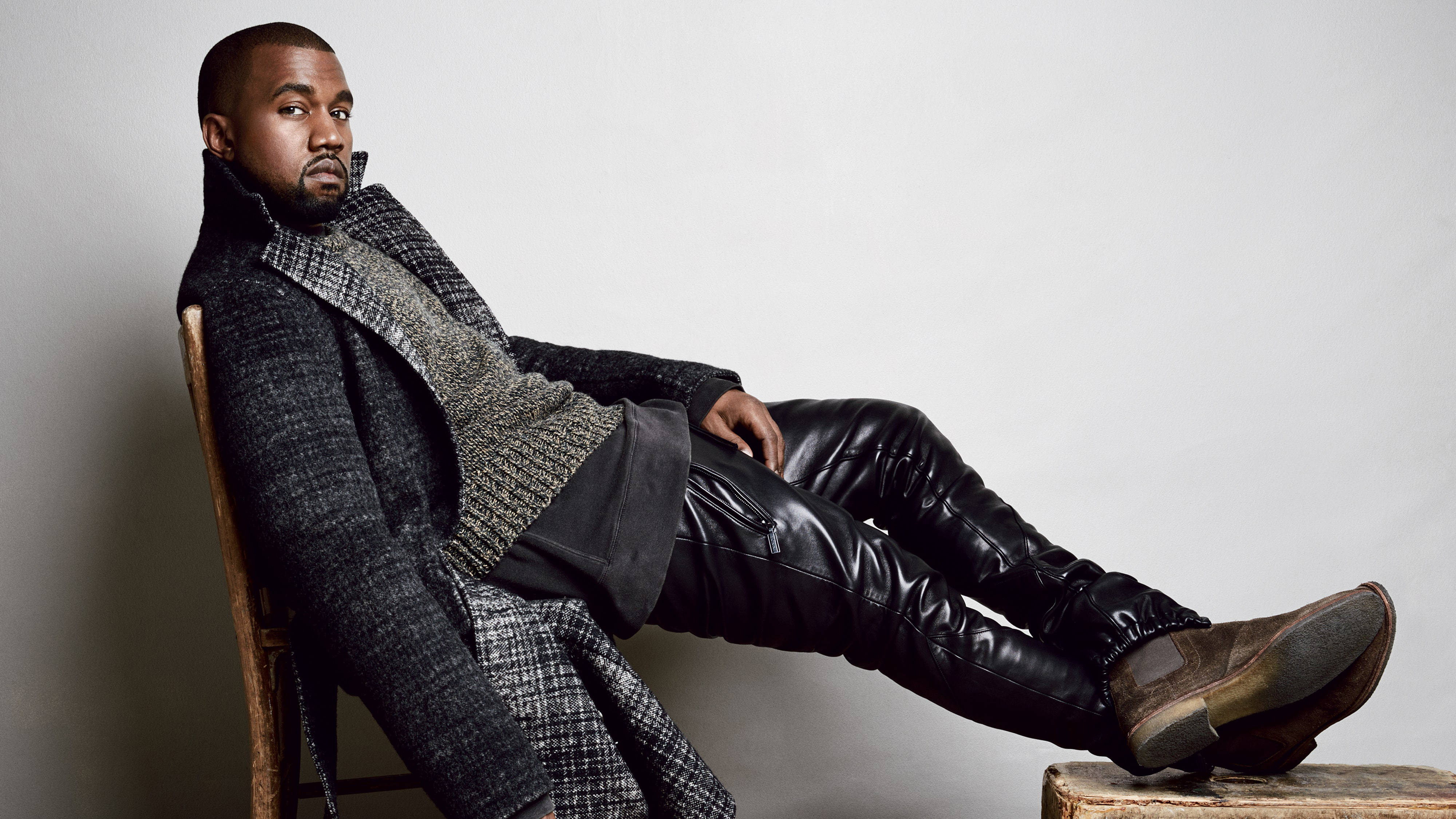 Kanye 桌布 壁紙 — Kanye West 自信地坐在椅子上，穿著質感外套和皮褲，背景是純灰色牆壁。
