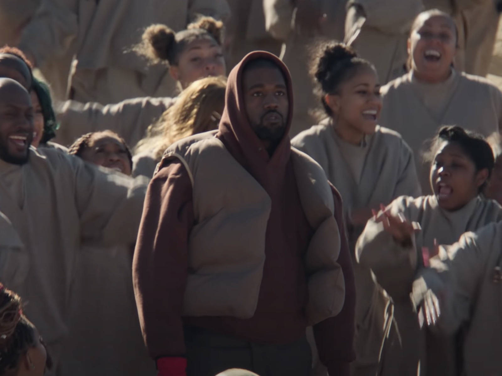 Kanye West Donda 桌布 壁紙 — Kanye West 身穿栗色連帽衫，在溫暖的陽光下，莊嚴地站在一群穿著米色長袍的歡樂人群中。