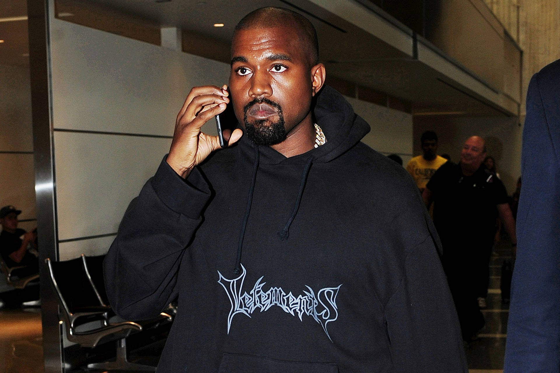 Vetements 桌布 壁紙 — Kanye West 身穿黑色連帽衫，上面有白色塗鴉風格文字，在現代室內走廊講電話。
