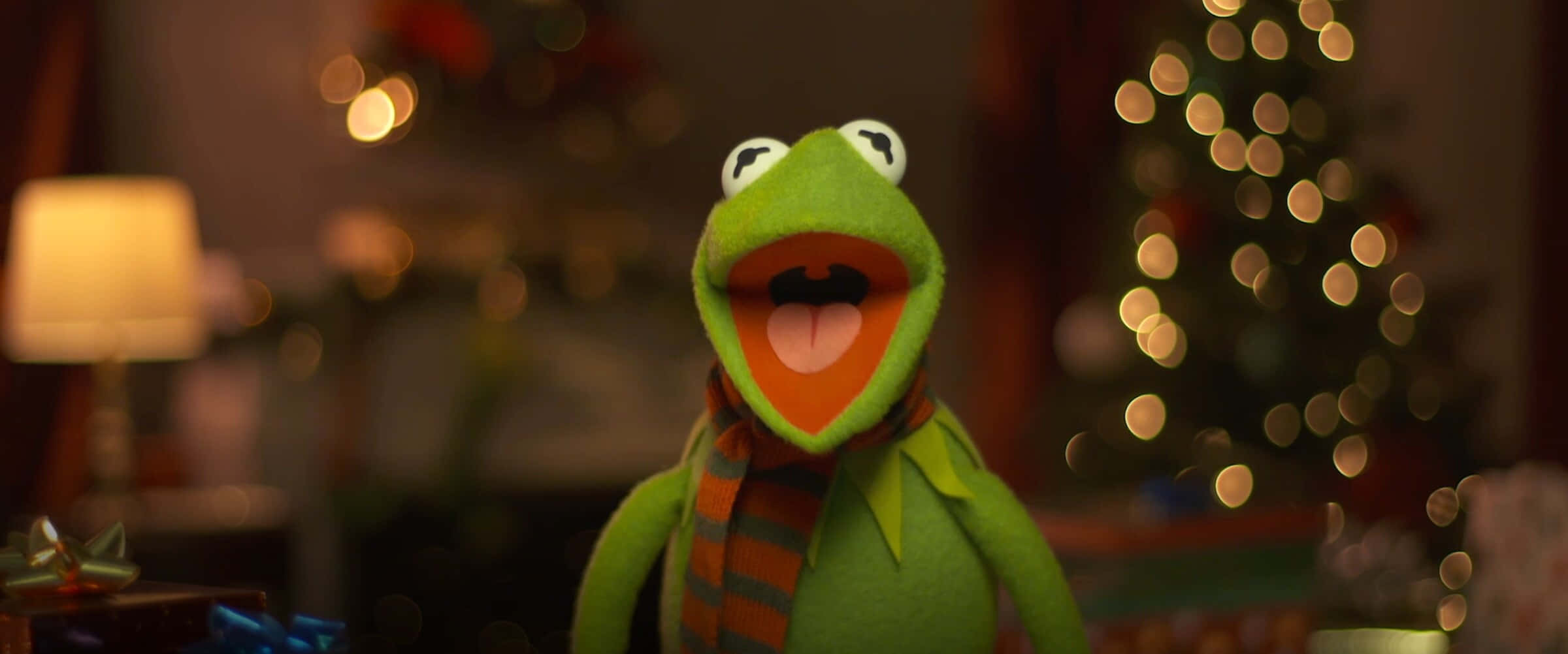Aesthetic Kermit 桌布 壁紙 — Kermit the Frog 戴著一條條紋圍巾，在一個溫暖照明的房間裡，背景有模糊的聖誕樹和燈，開心地微笑。