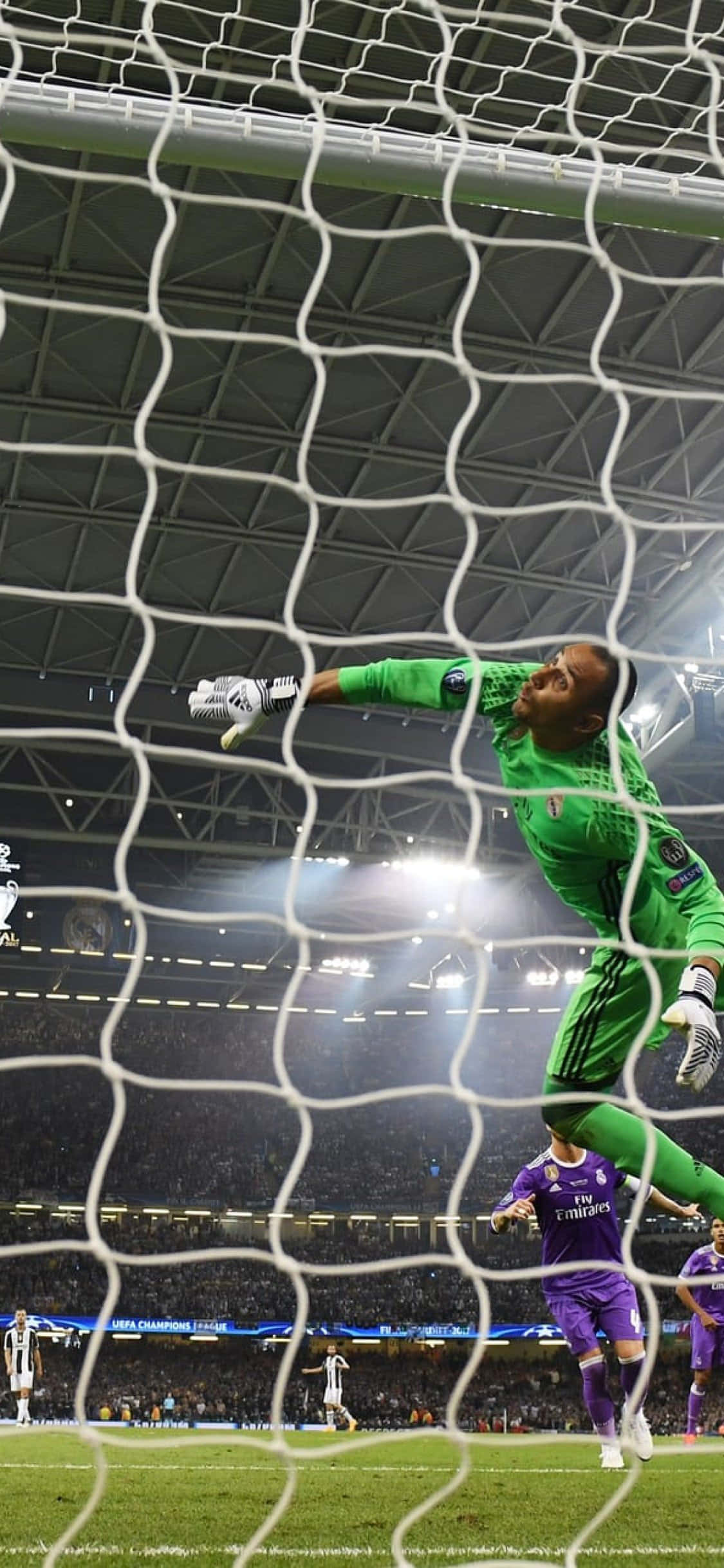 Keylor Navas 在球場上行動 壁紙 — 一名身穿綠色球衣的足球守門員在夜間比賽中跳過球網，在球場燈光下阻擋射門。
