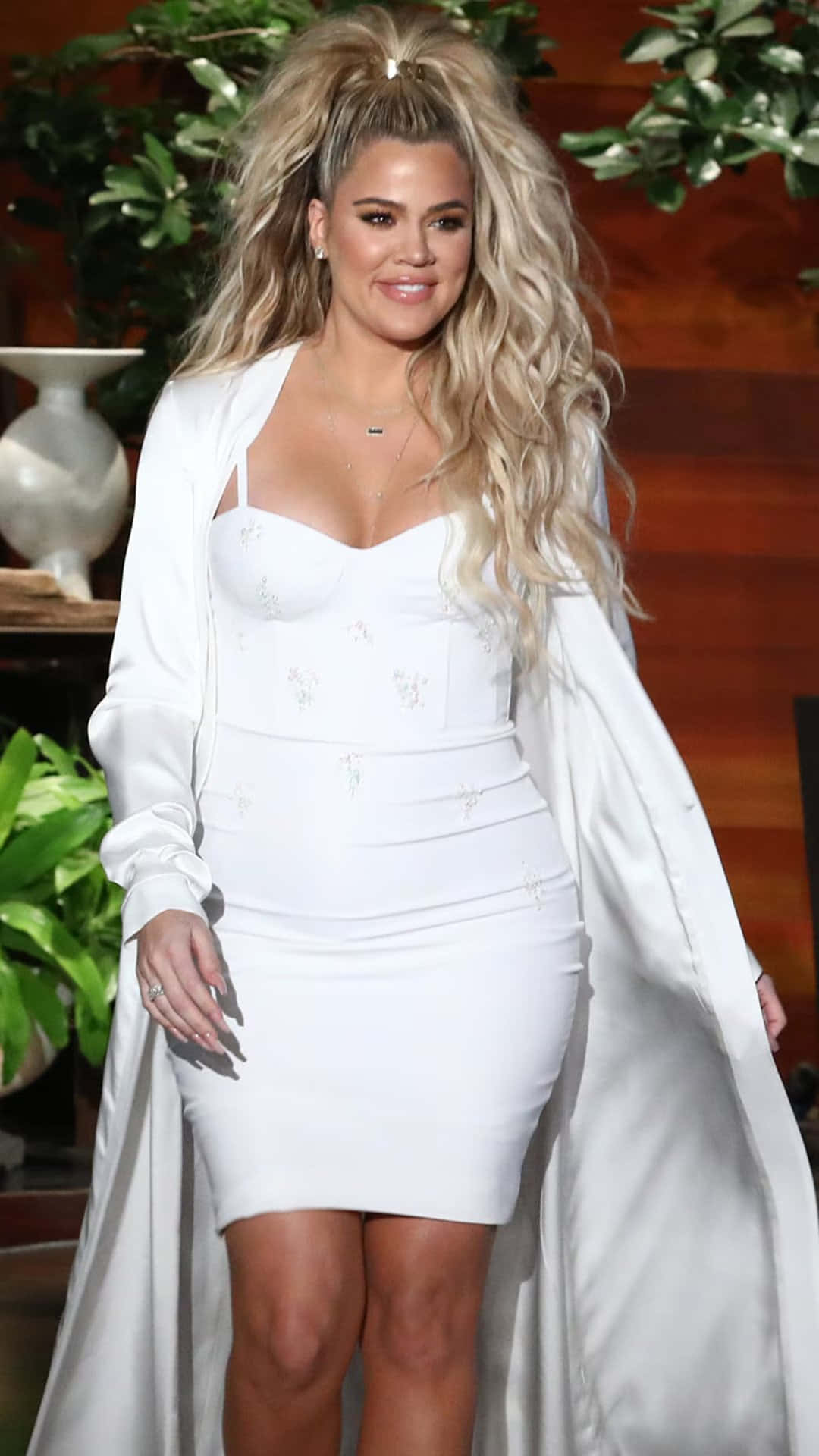 Khloe Kardashian 桌布 壁紙 — Khloé Kardashian 身穿白色貼身連衣裙和飄逸外套，一頭豐盈金髮，在木質背景和綠植襯托下微笑。