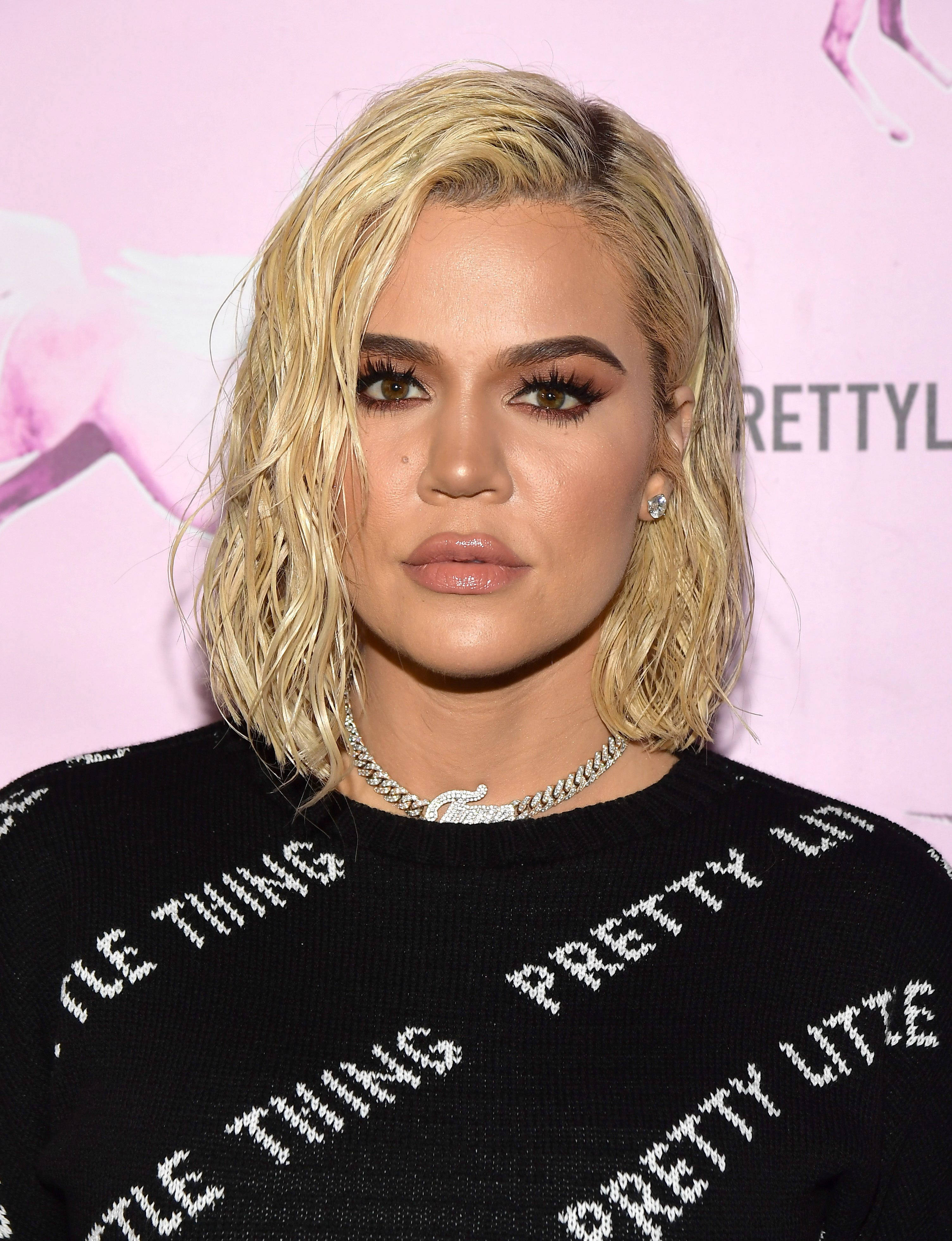 khloe 桌布 壁紙 — Khloé Kardashian 在粉紅色品牌背景前穿著帶有白色文字的黑色毛衣擺姿勢。