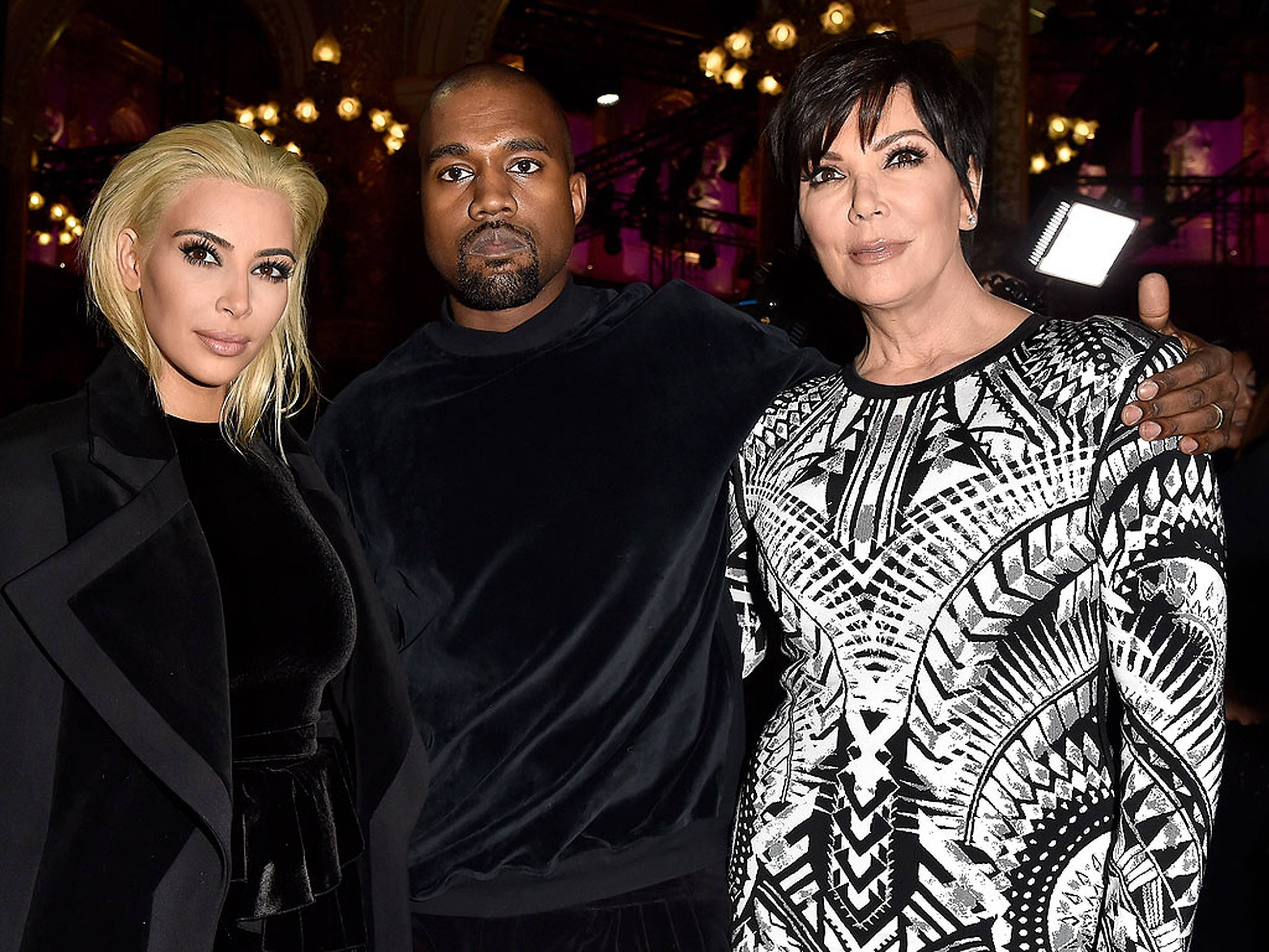 kris 桌布 壁紙 — Kim Kardashian、Kanye West 和 Kris Jenner 在一個燈光昏暗、裝飾華麗的吊燈和溫暖環境燈光的場地中一起擺姿勢。